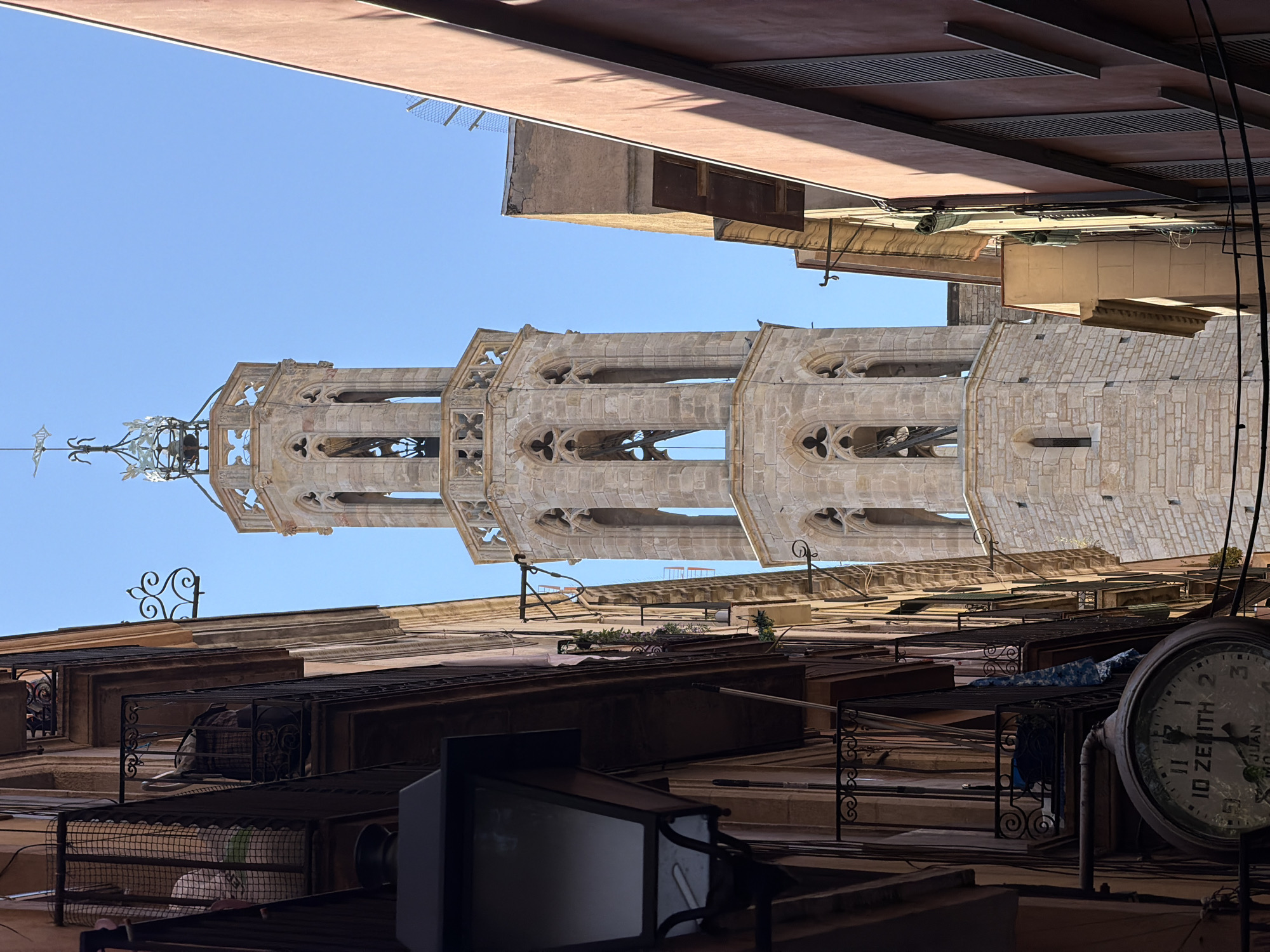 This is a vertical, eye-level medium shot of a tall, Gothic bell tower in Barcelona, Spain, viewed from a narrow alleyway. The tower, constructed of pale stone blocks, features several tiers with pointed arch windows and intricate tracery. The uppermost section of the tower is octagonal and capped with a decorative metal spire.
Flanking the tower are the sides of buildings. On the left, a dark, ornate lamp hangs in the foreground, partially obscuring a building facade with balconies. A large, old-fashioned clock face is visible on the lower left, showing the time as approximately 10:10. The building on the right is painted a reddish-orange and has closed shutters on its windows. The sky is a clear, bright blue. The scene captures the architecture of a historic European city, with a focus on the juxtaposition of the ancient bell tower and the surrounding urban environment.