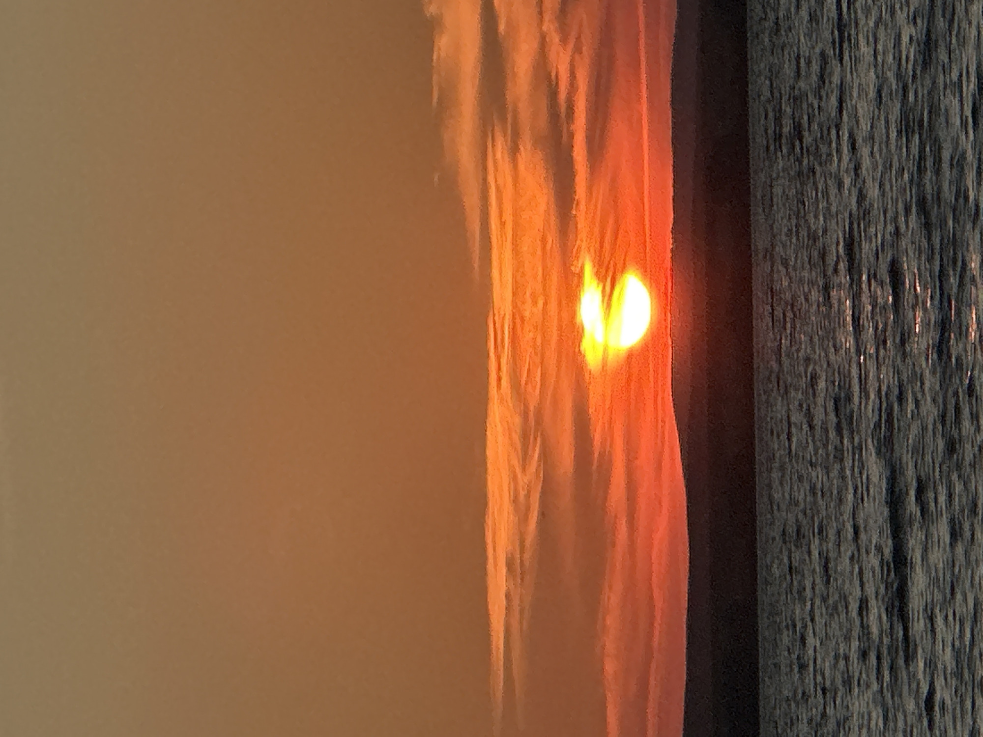 The image shows a sunset over a body of water, likely a lake or river, with a dark, silhouetted landmass in the background. The sky is a vibrant orange and red, with wispy clouds streaked across it. The sun, a bright yellow orb, is partially visible, casting a warm glow. The water is dark and choppy, reflecting the colors of the sunset. The scene is peaceful and serene. The city of Energetik, Russia, is located near the Zeya Reservoir, a large body of water known for its scenic beauty and sunsets. The image is consistent with a sunset view from this location. There are no people or man-made objects visible in the image, focusing solely on the natural landscape.