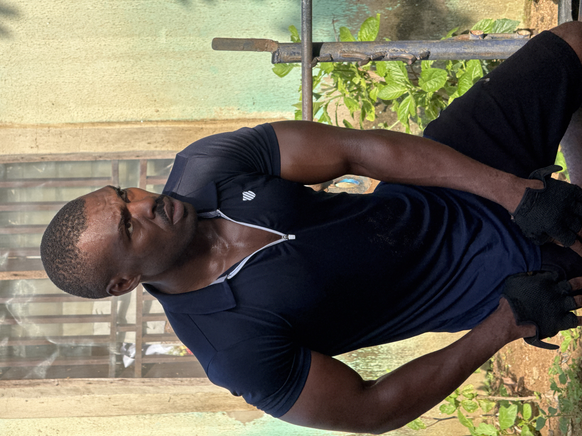 The image depicts a man with dark skin, a shaved head, and a mustache, wearing a dark blue short-sleeved polo shirt with a zipper and black athletic shorts. He is also wearing black fingerless gloves. He appears to be sweating, with his skin glistening. The man is positioned outdoors in what appears to be a somewhat dilapidated urban environment. Behind him is a building with a weathered, light green textured wall and a window with bars. To his left, there is a patch of dry, brown earth with sparse vegetation, and to his right, there are lush green plants growing around a rusty metal structure that resembles workout equipment. The overall impression is that of a person who has been engaged in physical activity outdoors. The presence of the building and vegetation suggests a warm climate, consistent with a location in Nigeria, specifically Akwanga.