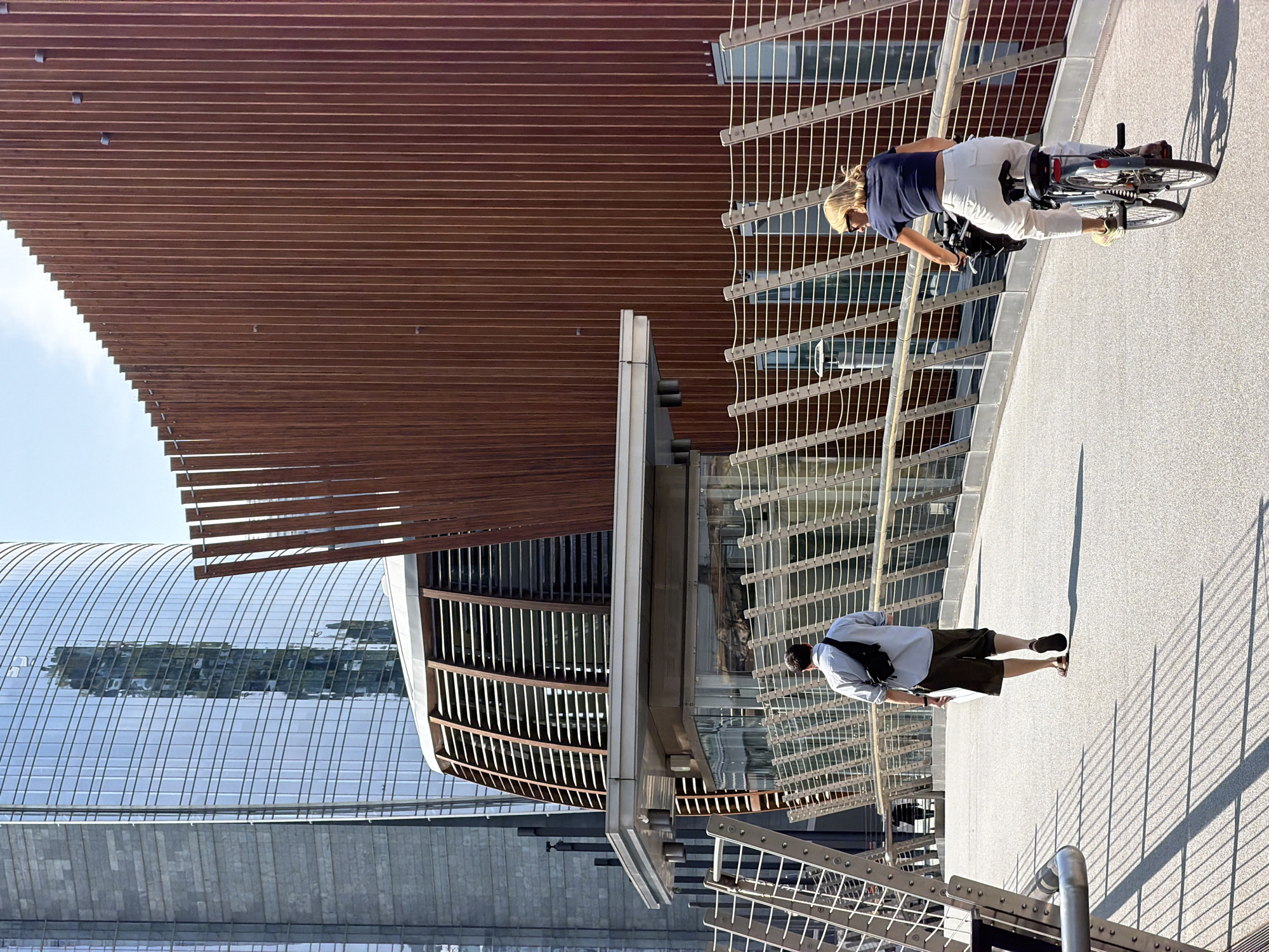 The image depicts a sunny outdoor scene on a pedestrian bridge in the Porta Nuova district of Milan, Italy. In the foreground, a man walks away from the camera, wearing a light blue collared shirt, khaki shorts, and sandals, with a black sling bag slung over his shoulder. To his right, a woman cycles away from the camera on a bicycle, wearing a dark blue t-shirt, white trousers, and sunglasses. She is viewed from behind, her blonde hair visible.
The bridge is characterized by a modern, light-colored railing system with diagonal white cables and metal supports, casting long, distinct shadows on the textured grey surface of the bridge.
In the background, contemporary architecture dominates the scene. To the left, a tall, slender skyscraper with a reflective glass facade is visible, showing reflections of other buildings and the sky. Next to it is a building with a grey, textured facade. In the center, a distinctive modern structure with a sloped roof covered in horizontal wooden louvers extends over the bridge. To the right, a large, reddish-brown building features a facade composed of vertical wooden slats. The sky is clear and blue. The overall impression is one of a modern urban environment with a focus on architectural design and pedestrian infrastructure.