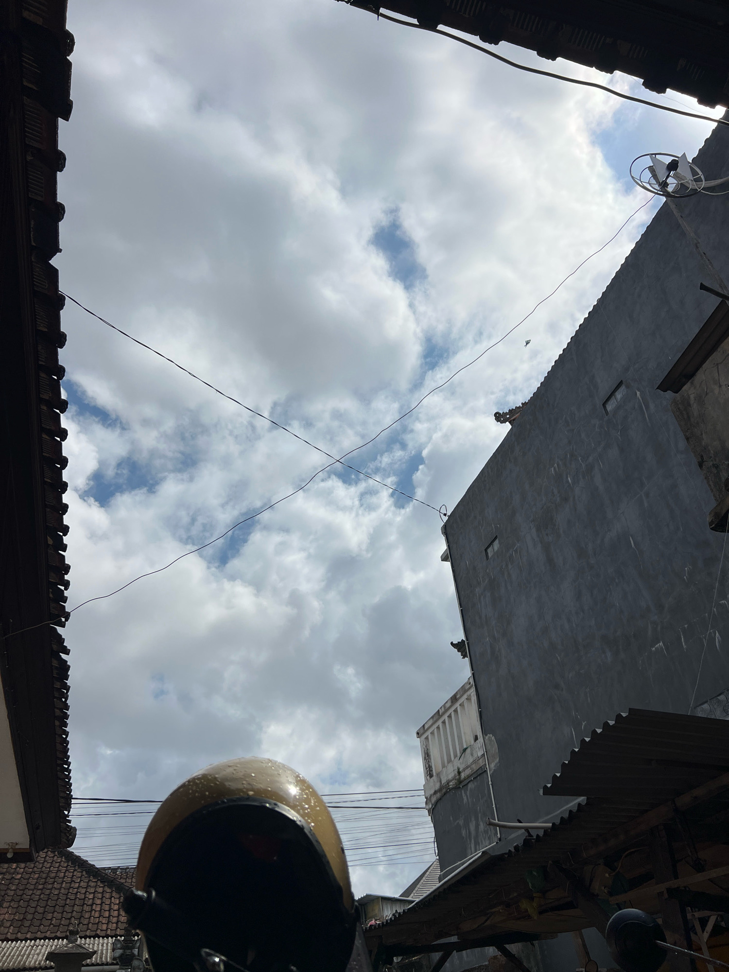 The image captures a low-angle view looking up at a cloudy sky in Denpasar, Indonesia. In the foreground, the top of a yellow motorcycle helmet is visible, its surface speckled with water droplets. On the right side of the frame, a dark gray, multi-story building dominates the view, with two small rectangular windows visible on its side. A satellite dish is mounted near the top right corner of the building. Several dark power lines crisscross the sky. On the left side of the image, a portion of a dark, tiled roof with decorative elements can be seen. The overall atmosphere suggests a humid or recently rained-on environment.