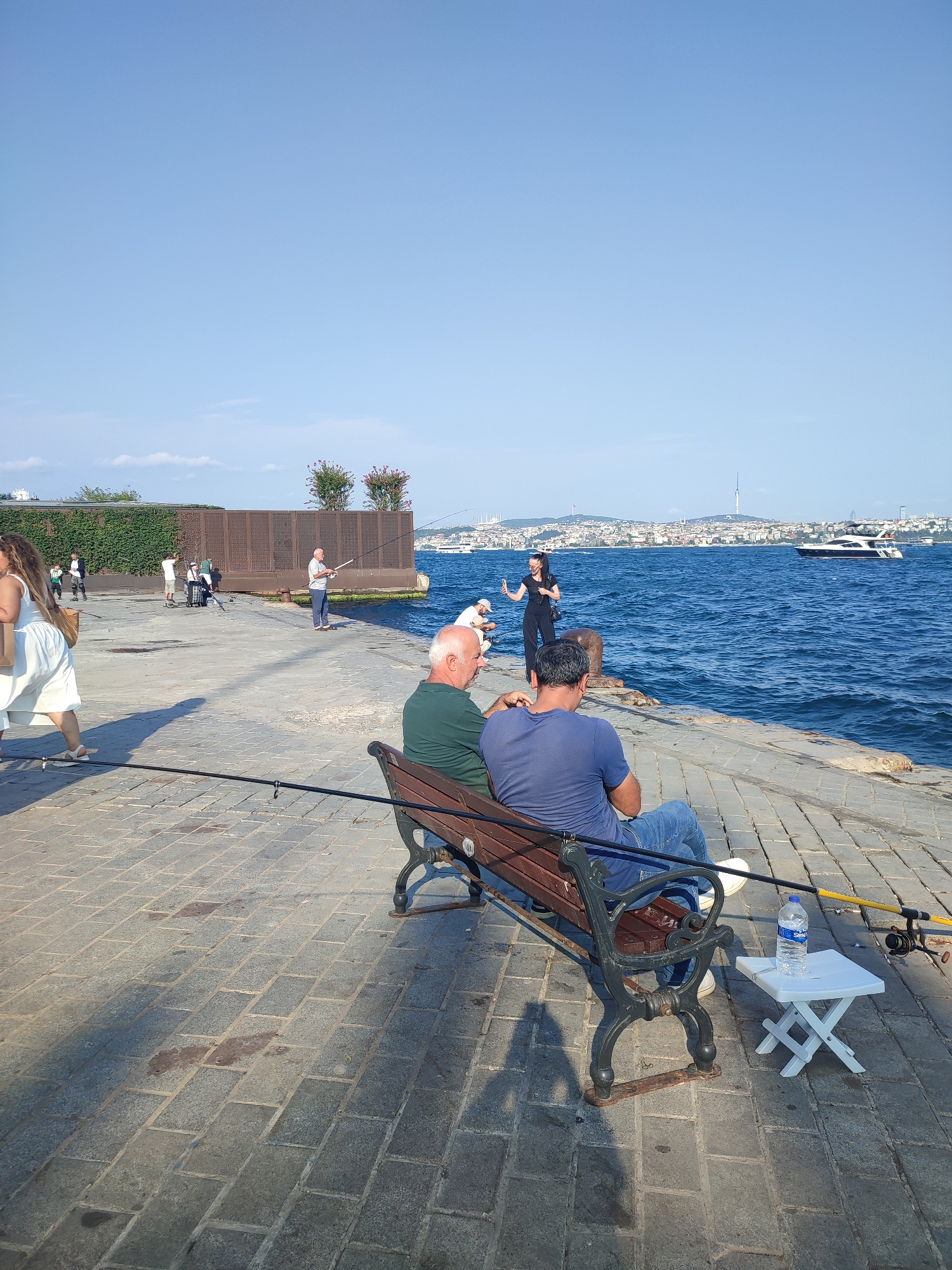 In Beyoğlu, Turkey, on a sunny day, two men are seated on a park bench facing the sea, engaged in fishing. The man on the left wears a green polo shirt and the man on the right wears a blue t-shirt and jeans. Two fishing rods are set up beside them, with one extending onto the bench. To the right of the bench, a small white foldable table holds a bottle of water. The paved walkway extends to the left where a woman in a white dress walks away from the camera, carrying a shopping bag. Further left, several other individuals are seen fishing or walking along the waterfront. In the distance, a cityscape with buildings and a tower is visible across the water, and a white yacht sails by. The sky is clear blue.