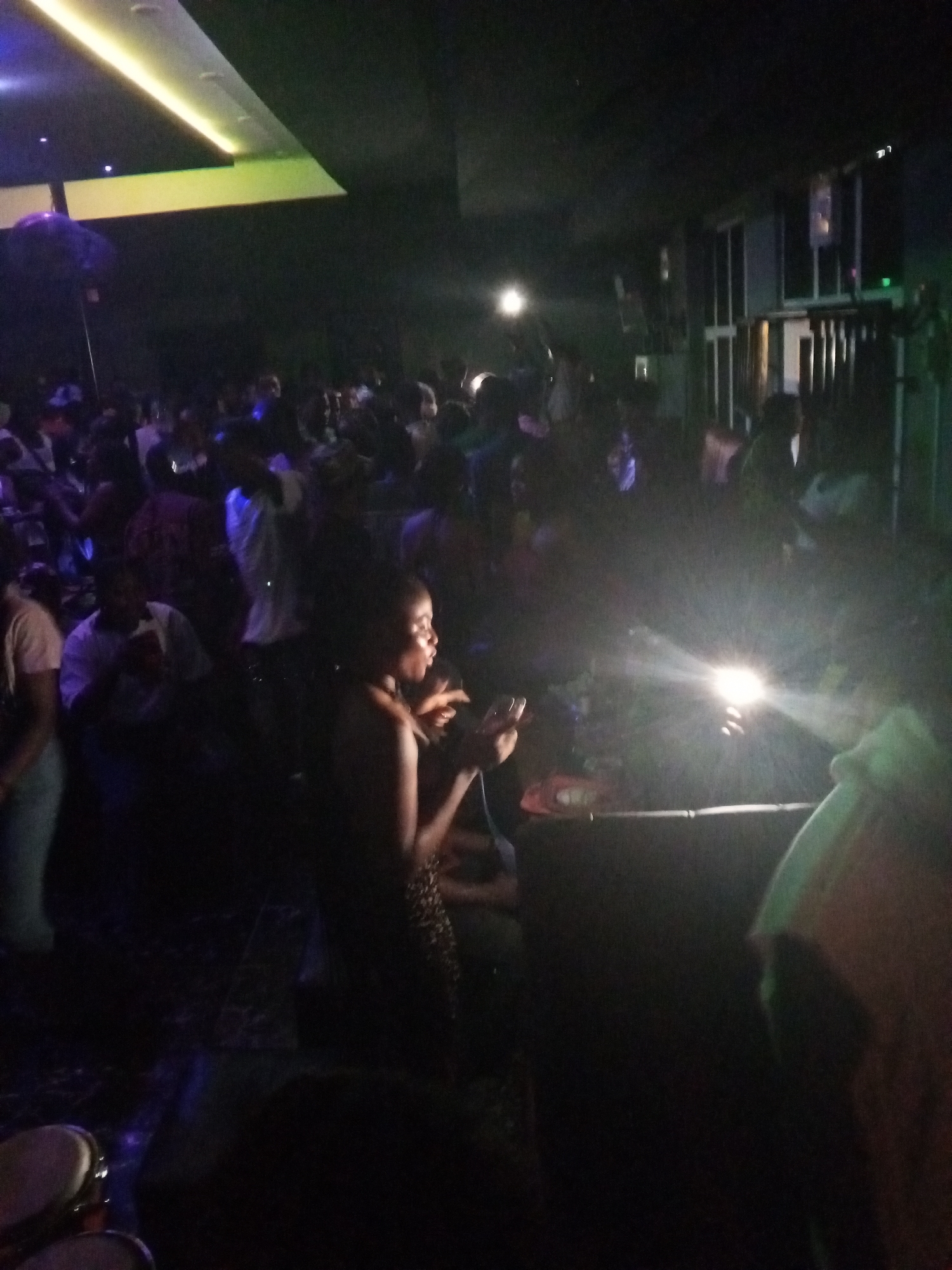 A dimly lit indoor scene depicts a crowded space, possibly a nightclub or a gathering in Lokoja, Nigeria. Numerous individuals are present, with one woman in the foreground appearing to be speaking or singing, possibly holding a microphone. The lighting is low, punctuated by brighter spotlights that create a sense of depth and highlight certain areas. The background is filled with indistinct figures, suggesting a social event. Some musical instruments are also partially visible.