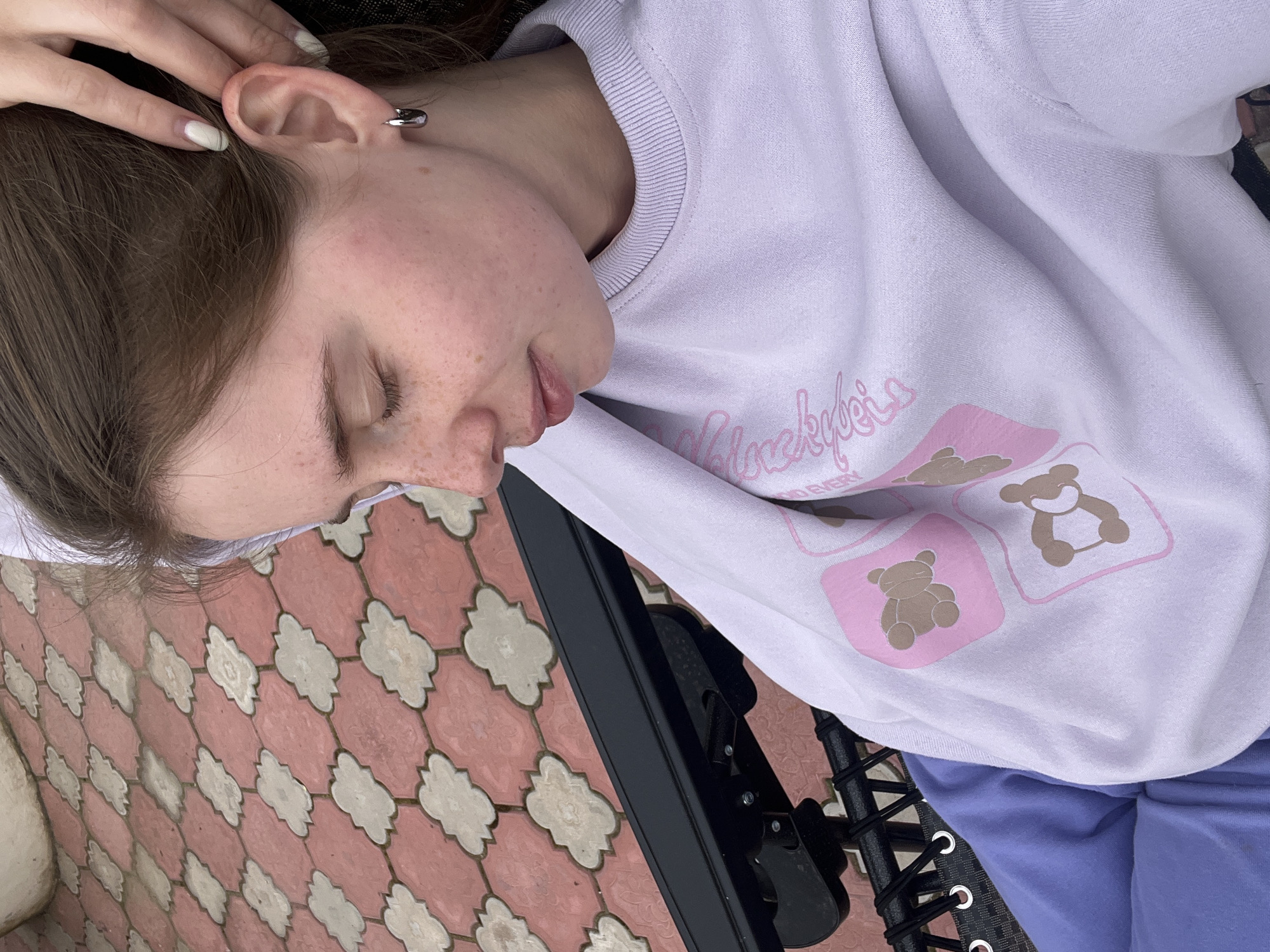 A young woman is lying down, possibly sleeping, outdoors. Her head rests on a white surface, perhaps a cushion or pillow. She is wearing a light-colored sweatshirt with teddy bear designs and light blue pants. Part of a black metal frame, possibly from a piece of furniture or equipment, is visible beneath her. The background consists of patterned red and white paving stones. The location is Chaikovsky, Russia.