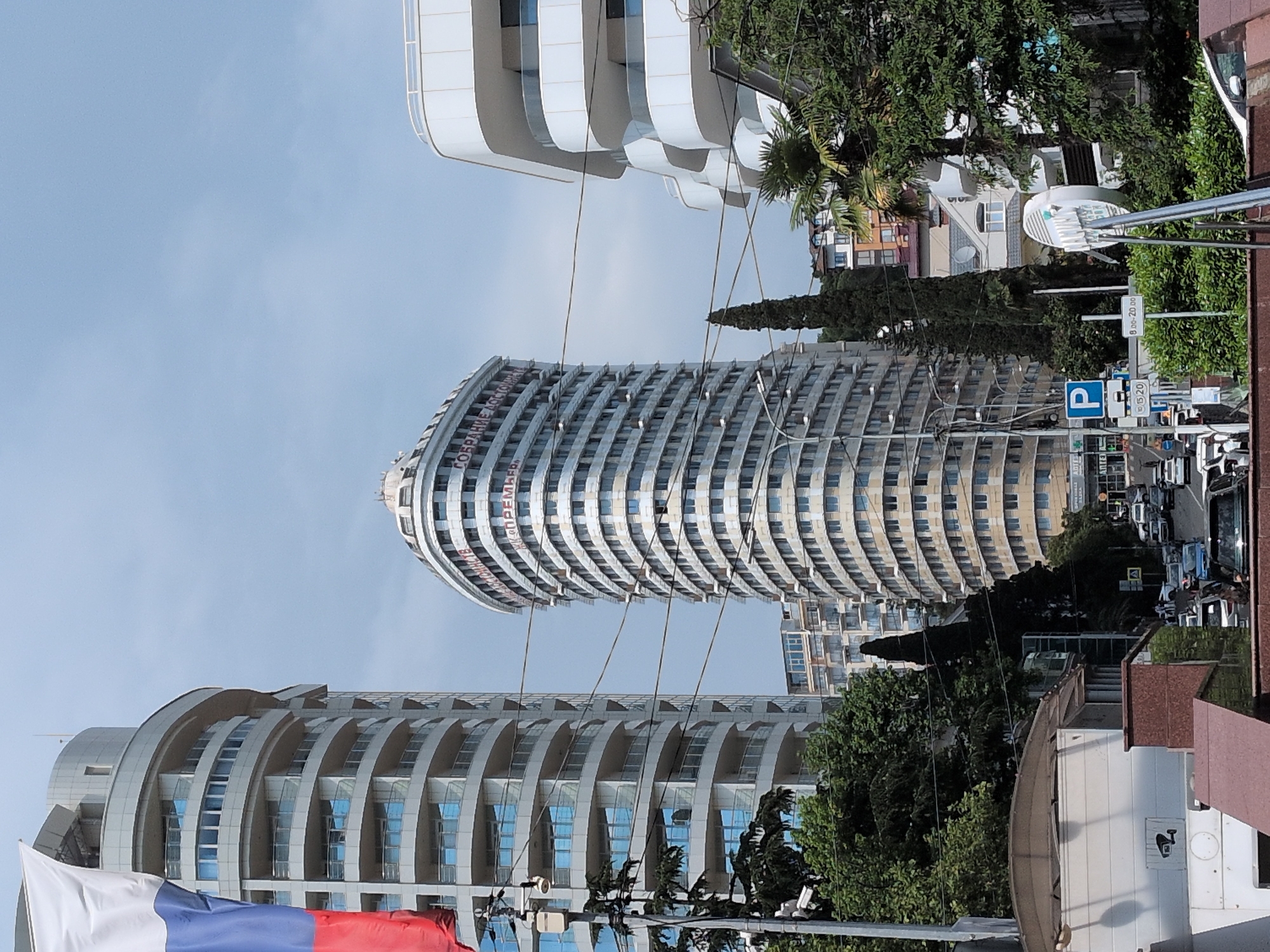 This is a photograph of the coastal city of Sochi, Russia, on a cloudy day. The scene features a prominent, curved building with multiple stories and balconies. The building's facade is a light color, and it has a distinctive, almost boat-like shape. Above the building, a Russian flag is visible, attached to a pole. In the foreground, a street is visible with several cars, and some greenery, including trees, is present. Other buildings of varying heights and architectural styles are also in the background, contributing to the urban landscape. A traffic sign with a "P" indicating parking is also visible. The image captures a street-level view of the city, with modern architecture and everyday urban elements.
