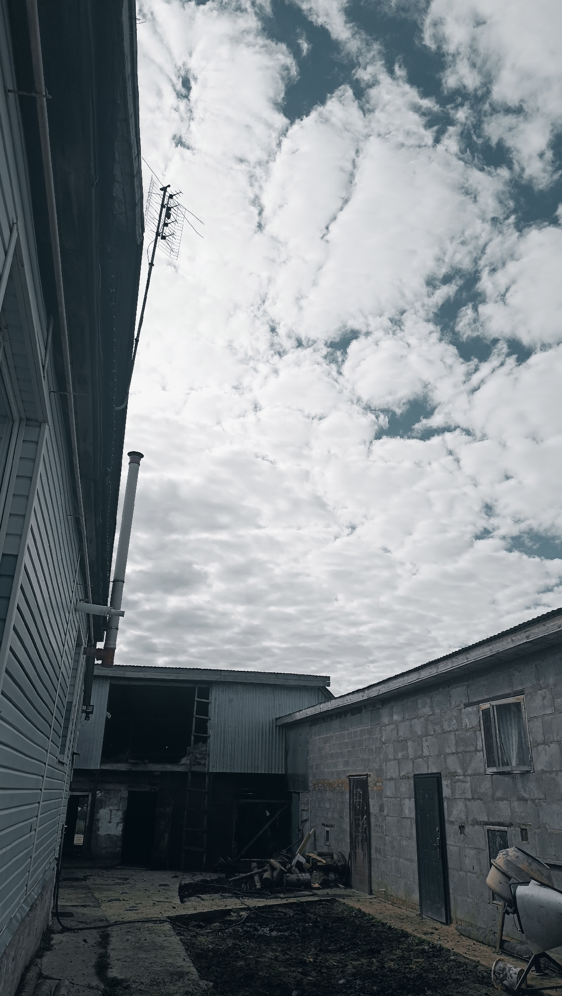 The image depicts an outdoor scene on a cloudy day, with a low angle perspective looking upwards. In the foreground, to the left, is the side of a building with light-colored horizontal siding and a dark roofline. A white pipe with red bands extends upwards from this building. To the right of this building, further into the scene, are other structures. One appears to be a corrugated metal building with a large open doorway, and a ladder leaning against it. Adjacent to this, to the right, is a building constructed of gray cinder blocks. This building has a few doors and at least one window with curtains. In front of the cinder block building, a concrete mixer is partially visible. The ground in the foreground and midground is dark and appears to be muddy or soil. There are also some logs and debris scattered on the ground. In the background, the sky is filled with numerous white and gray clouds against a dark blue sky. An antenna is visible attached to a pole extending from the side of the building on the far left. The overall impression is of an industrial or utilitarian area, possibly a farm or workshop, in a somewhat rundown state. No people are visible in the image. The image does not provide any specific visual cues that would allow for localization to Karsha, Russia.