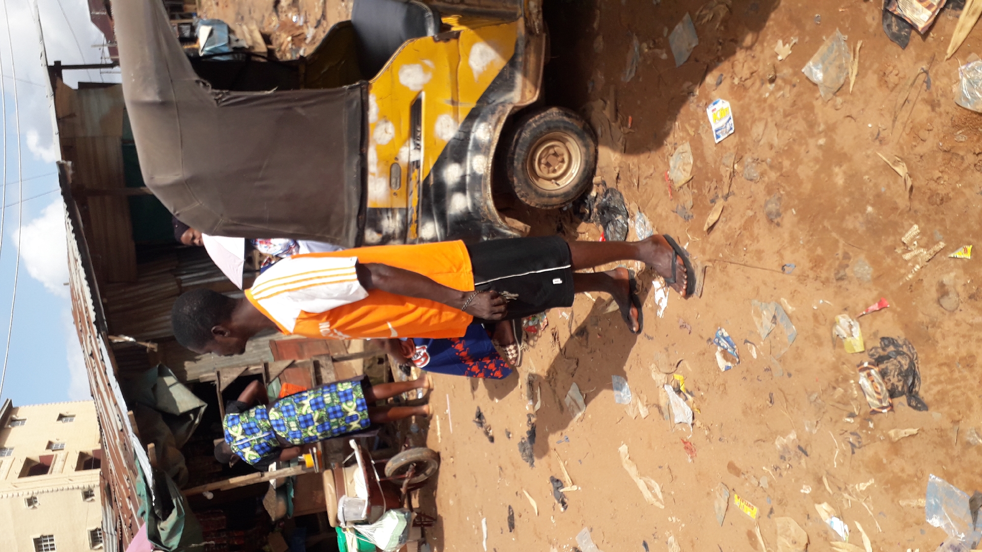 In Nkpor, Nigeria, a man stands in front of a yellow and black auto-rickshaw on a dirt road littered with debris. He wears an orange and white t-shirt and black shorts. Behind him, a woman in a blue and green patterned dress stands near a market stall. Another person, partially visible, holds a pink umbrella. The scene suggests a bustling market or roadside area.