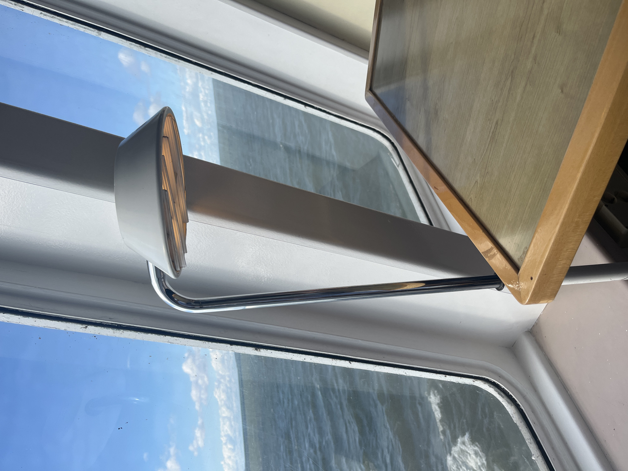 The image depicts an interior view from a window, likely on a ferry or boat, with a view of the sea under a partly cloudy sky. In the foreground, a wooden table with a light brown laminate top and a wooden frame is visible on the right. A chrome lamp with a white conical shade extends from the left, positioned above the table. Through the window, the dark blue sea with white-capped waves is visible, and a strip of distant land can be seen on the horizon. The interior framing of the window is white. The scene is devoid of people. The location is identified as Virtsu, Estonia.