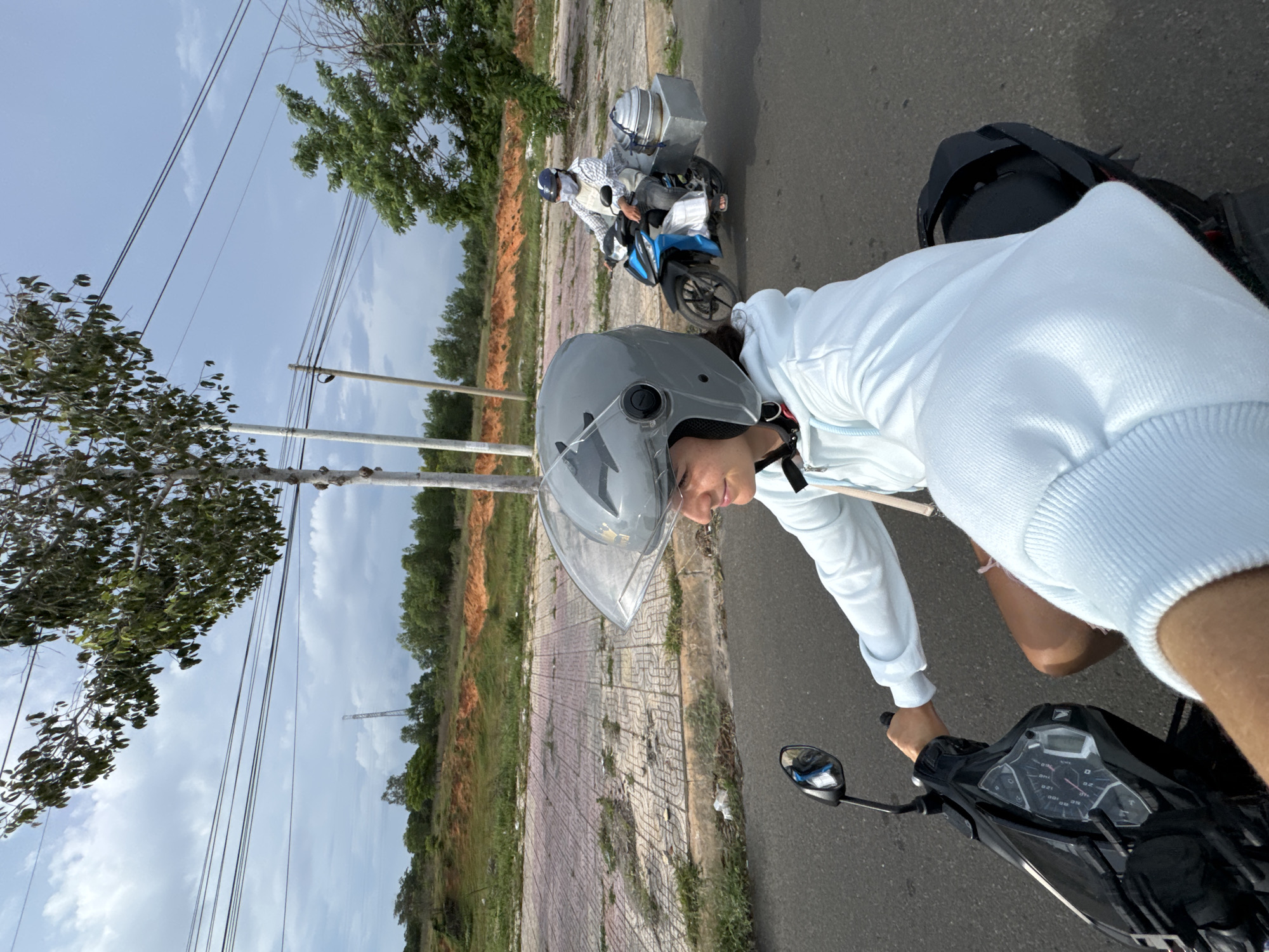 Two people are riding motorbikes on a paved road in Phan Thiết, Vietnam. In the foreground, a woman wearing a light blue hooded sweatshirt and a gray helmet is riding a black motorbike. She is looking to her right with a slight smile. Her hands are on the handlebars, and the motorbike's dashboard is visible. In the background, a second person wearing a helmet, a checkered shirt, and a white apron is riding a blue motorbike. This motorbike has a metal cargo box attached to the back containing two stacked metal containers. The environment includes a sidewalk with patterned tiles, green grass, red dirt hills, trees, and power lines overhead. The sky is partly cloudy.