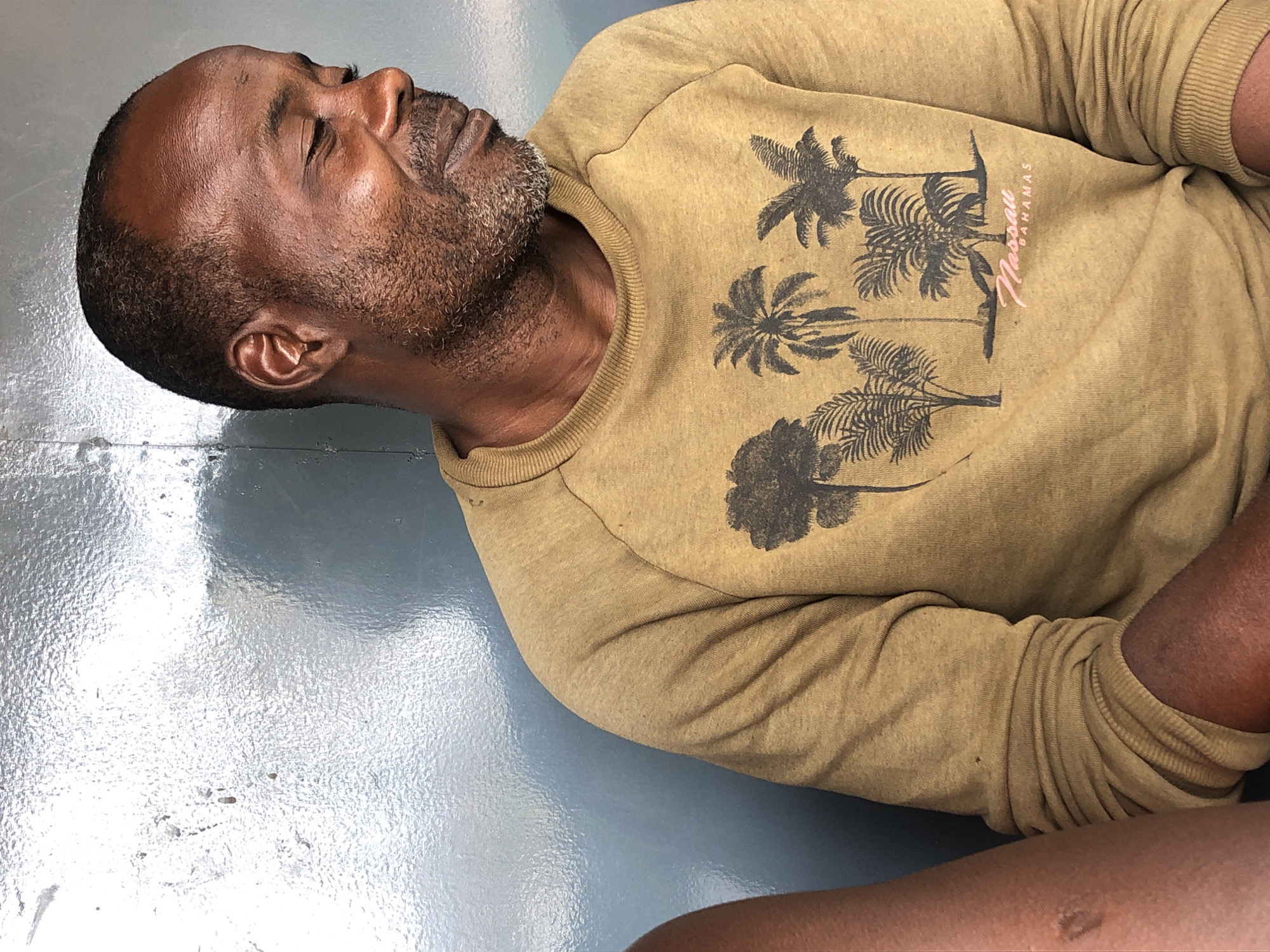 In Jos, Nigeria, a man of African descent with short hair and a beard is captured in a medium shot, looking down and to the right with his eyes closed. He is wearing a mustard-colored sweatshirt with a graphic of palm trees and the word "Nassau Bahamas" printed on it. His right arm is visible, resting on what appears to be his lap. To his left, a portion of another person's arm, with lighter skin tone and a scar, is visible. The background is a textured, greyish-blue wall with a reflective sheen, possibly made of metal or a similar material. The lighting is soft, creating a gentle sheen on the man's face and highlighting the texture of the background.