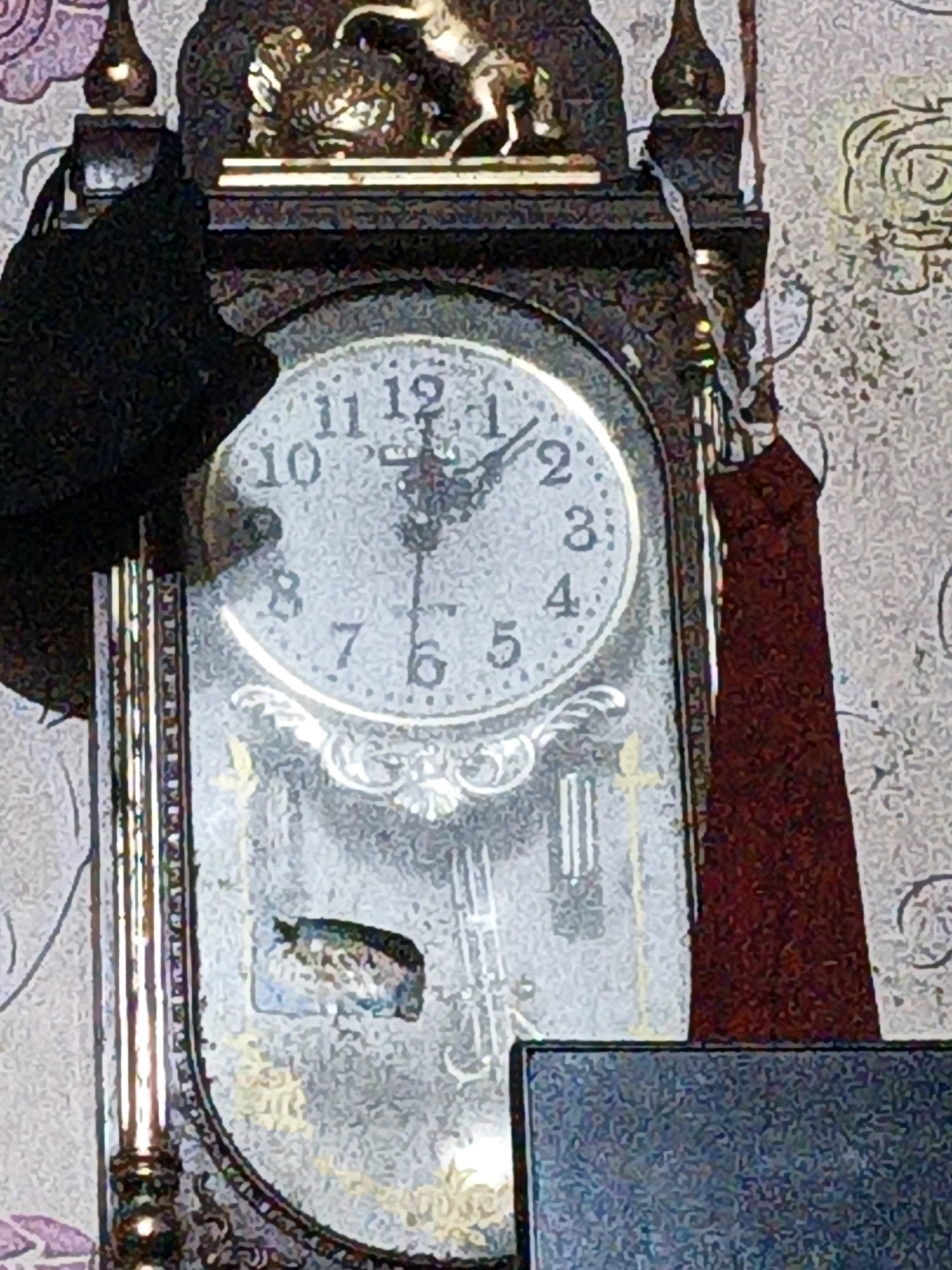 This is a close-up, slightly blurry, indoor photograph of the face of an antique pendulum clock. The clock has Roman numerals indicating the hours, and two hands are visible. The hour hand appears to be pointing towards the 11, and the minute hand is pointing towards the 1. The clock face has a decorative, ornate design. The clock is dark brown and appears to be made of wood. A dark fabric, possibly a curtain or cloth, is draped over the left side of the clock, and a dark red fabric is draped over the right side. The background is a textured wallpaper with a floral or ornate pattern. The image does not provide any contextual information about the city of West Jakarta, Indonesia, or any specific location within it.