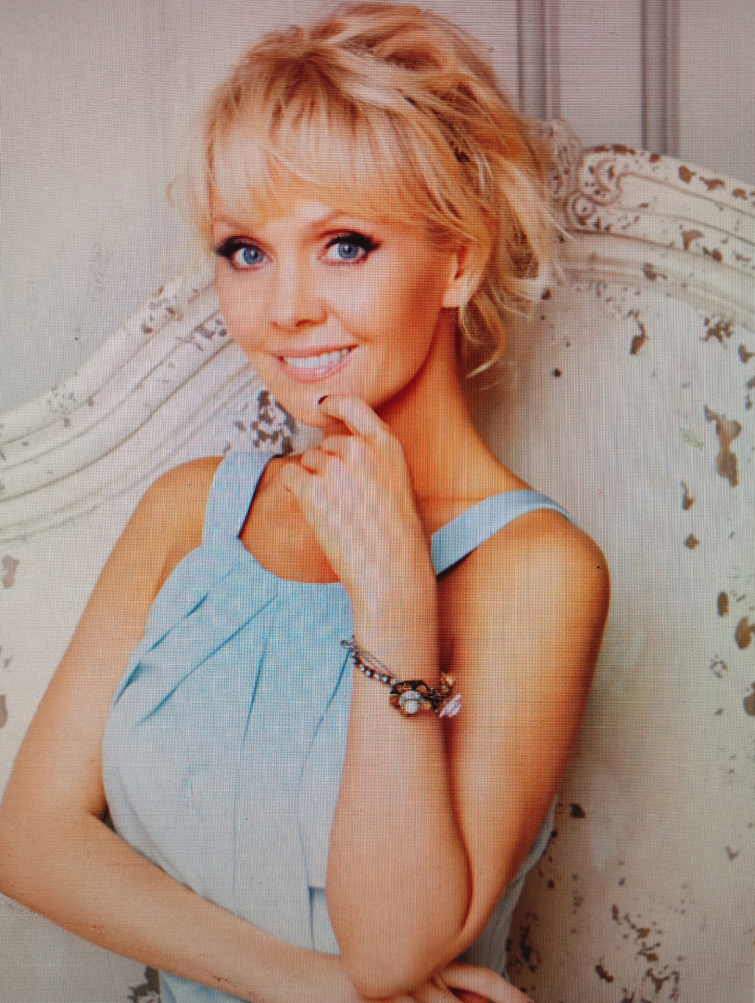 A woman with blonde hair and blue eyes, wearing a light blue top, poses with her hand on her chin. She is seated on a distressed white chair with ornate carvings. The background is a light-colored wall with an aged texture. The image is a medium shot, focusing on the woman's upper body and face. The overall scene is softly lit, creating a gentle and inviting atmosphere. This scene does not provide any information to determine its location in the city of Sluck, Belarus.