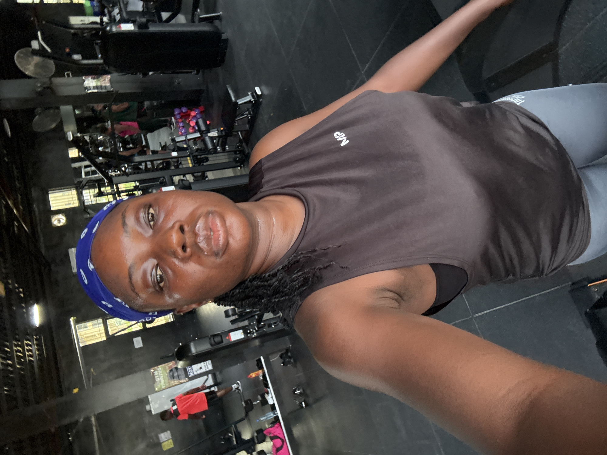 A woman with dark skin and braids wears a blue headscarf and a dark tank top with the "MP" logo. She is in a gym setting, with exercise equipment visible in the background, including weight machines and dumbbells. The environment appears to be dimly lit, with concrete walls and floors. Other gym-goers are visible in the background, including a person in a red shirt walking away from the camera. The overall scene suggests a workout session in progress at a gym in the city of Iddo Tudunwada, Nigeria.