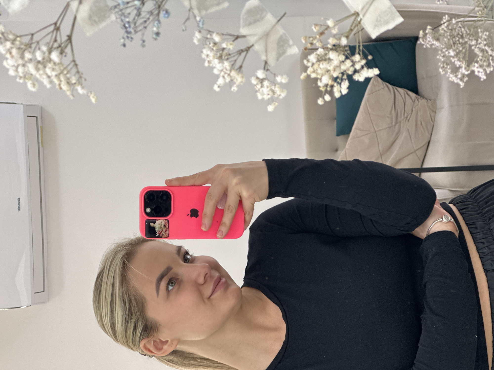 A woman with blonde hair takes a selfie using a pink phone case. The photo appears to be taken in a room with a mirror. Decorations of baby's breath flowers are above her, and in the background, pillows and a sofa can be seen. An air conditioning unit is on the left wall. The specific location in Budva, Montenegro cannot be determined from the image.