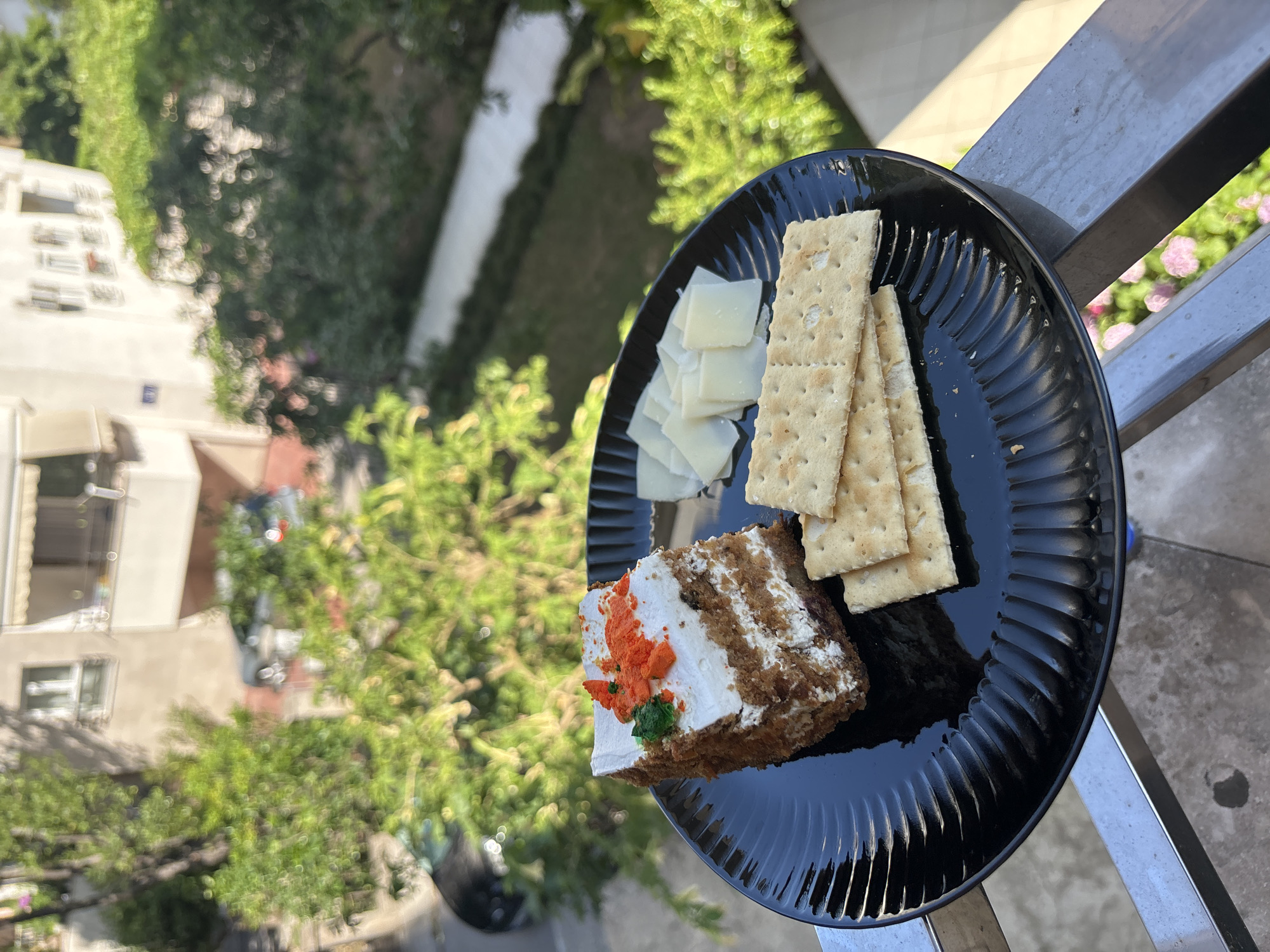 A slice of carrot cake topped with white frosting and orange and green sprinkles sits on a black ridged plate. To the right of the cake are several slices of white cheese and three crackers. The plate is positioned on a railing overlooking a street and building. Green trees and a parked car are visible in the background. The image appears to have been taken on a balcony in Budva, Montenegro, during daylight.