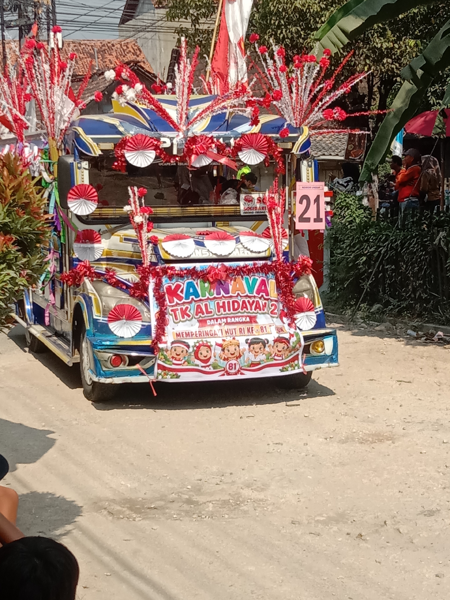 The image shows a decorated vehicle, likely a jeep or a similar open-top vehicle, participating in a parade or festival in Kunden, Indonesia. The vehicle is adorned with red and white decorations, including streamers, fans, and floral arrangements, in patriotic colors. A banner on the front of the vehicle reads "KARNAVAL TK AL HIDAYAH 2" and "MEMPERINGATI HUT RI KE-81", indicating it is a carnival parade for the Al Hidayah Kindergarten, celebrating Indonesia's 81st Independence Day. Several children are depicted on the banner. A large sign with the number "21" is visible on a post to the right of the vehicle, possibly indicating a route number or participant number. In the background, there are residential buildings with tiled roofs, trees, and a few onlookers dressed in casual attire. The ground is unpaved, suggesting a street in a village or less urbanized area. The lighting suggests it is daytime, with sunlight casting shadows.