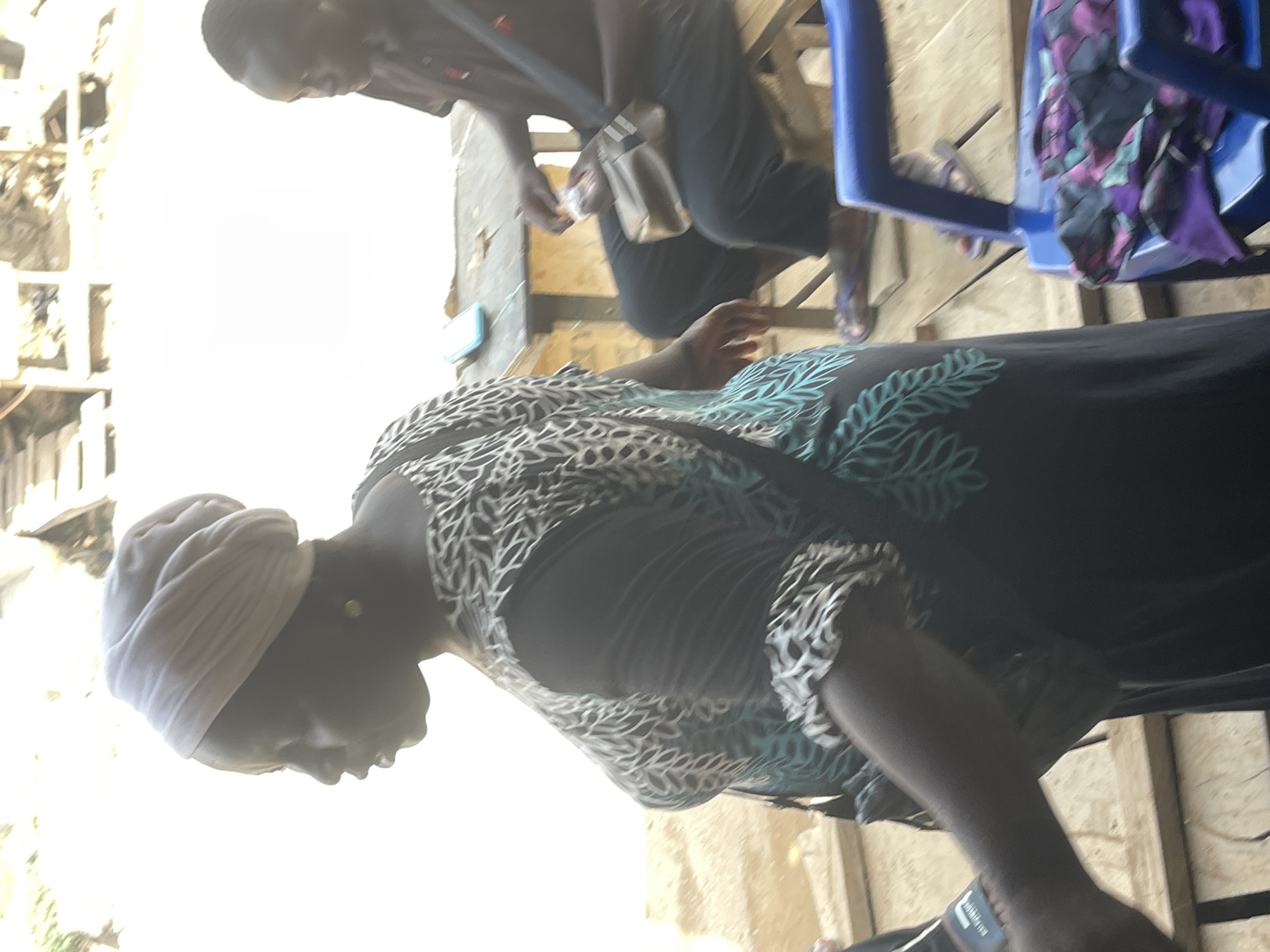 The image shows a woman in a printed dress and head wrap, possibly outdoors in Abuja, Nigeria, based on general context. Another person is visible in the background seated near a blue chair. The focus is on the woman and the immediate surrounding area.