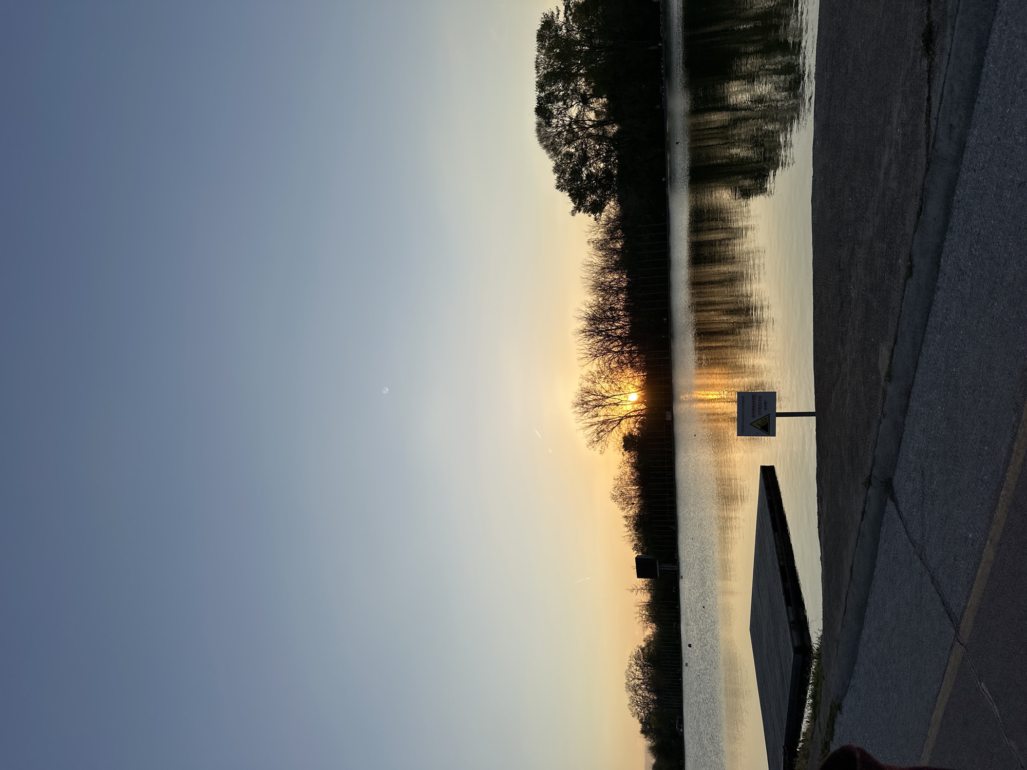 The image captures a tranquil scene in Plovdiv, Bulgaria, likely near a body of water such as a lake or river. The sun is setting, casting a golden glow through silhouetted trees along the water's edge, which create reflected image in the still water. There is a concrete pathway or embankment to the right side of the image. A warning sign is visible at the right. The sky is clear with a color gradient, ranging from a soft blue to a warm golden hue near the horizon.