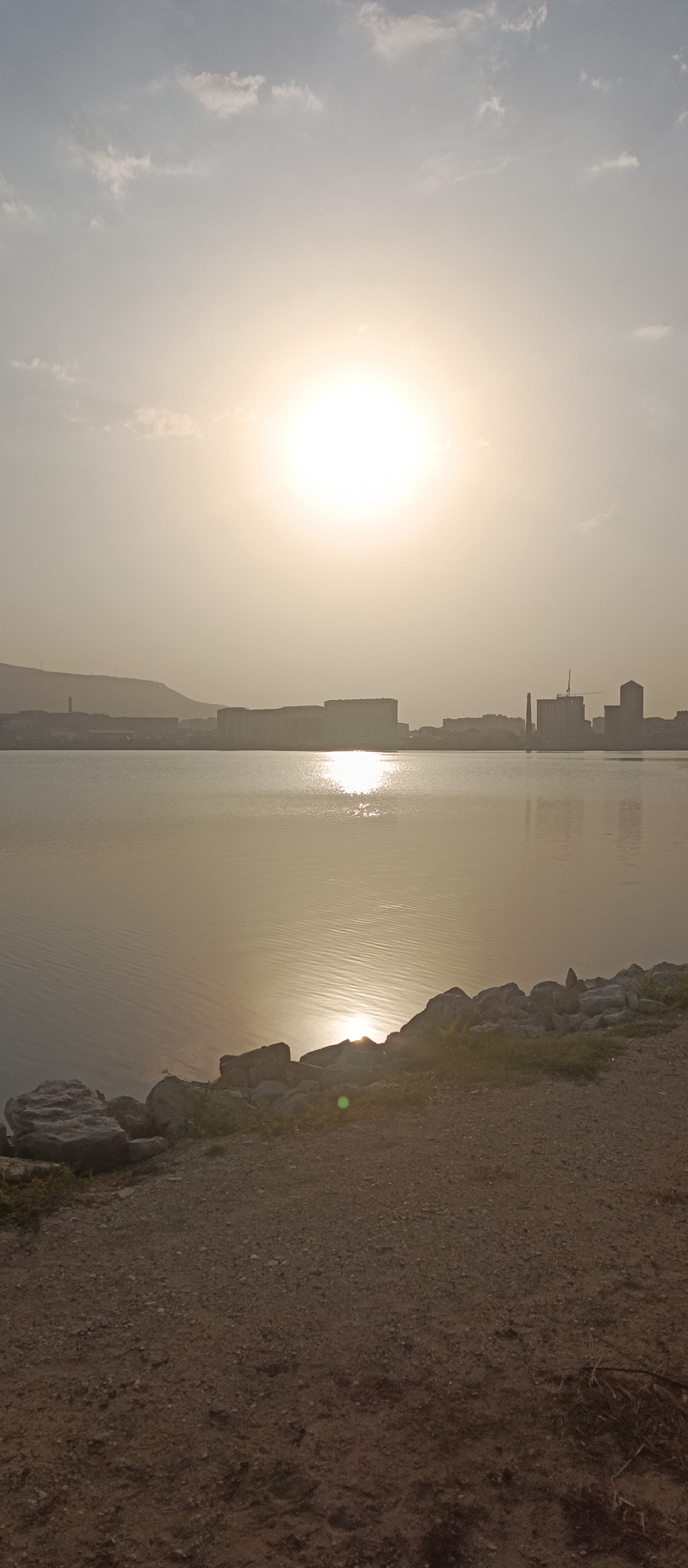 This is a vertical, eye-level shot of a tranquil bay in Makhachkala, Russia, under a hazy, bright sky with the sun prominently visible. The sun's glare creates a shimmering reflection on the calm water, which dominates the mid-ground of the image. Along the distant shoreline, a silhouette of the city is discernible, featuring low-rise buildings, industrial structures like a chimney, and a mountain range in the background, partially obscured by haze. The foreground consists of a rocky embankment transitioning into a gravelly path. There are no people or animals present in the scene.