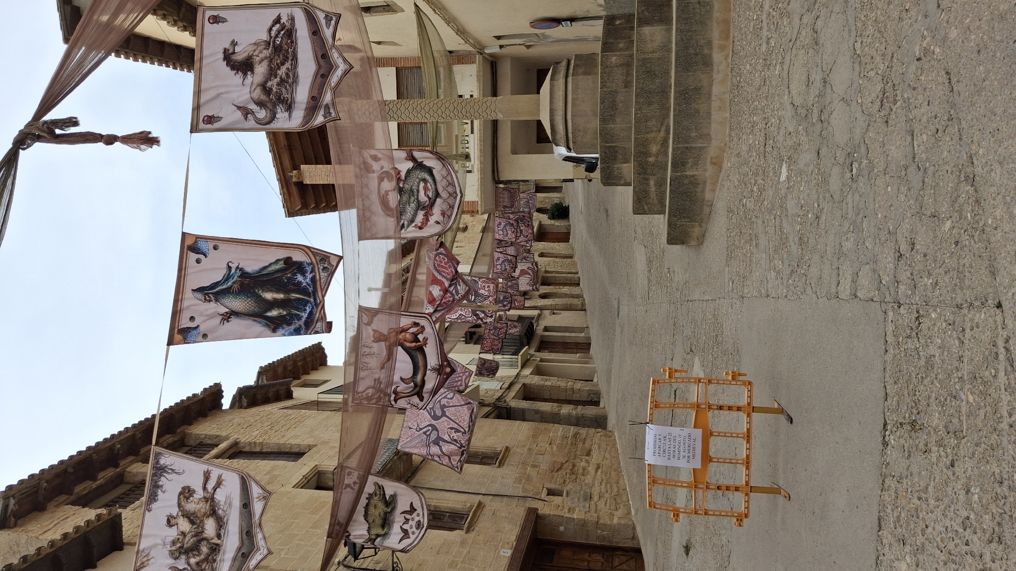 The image depicts a narrow street in the town of La Fresneda, Spain, decorated with numerous medieval-themed banners hanging from overhead wires. The banners feature various illustrations, including mythical creatures, animals, and human figures, some of which appear to be related to heraldry or folklore. The street itself is paved with cobblestones and concrete, showing signs of age and wear. Buildings with stone facades and tiled roofs line both sides of the street, suggesting a historic urban setting. In the foreground, an orange plastic barrier is placed on the street, with a sign attached that reads in Spanish "PROHIBIDO APARCAR Y CIRCULAR HASTA LAS 21 HORAS DEL DOMINGO, 16 DE AGOSTO POR MERCADO MEDIEVAL", indicating a prohibition on parking and circulation until 9 PM on Sunday, August 16th, due to a medieval market. To the right of the street, a stone monument with tiered steps stands prominently. The sky is overcast, contributing to a subdued lighting condition. The scene appears to be set up for or anticipating a local event, likely the medieval market mentioned on the sign.