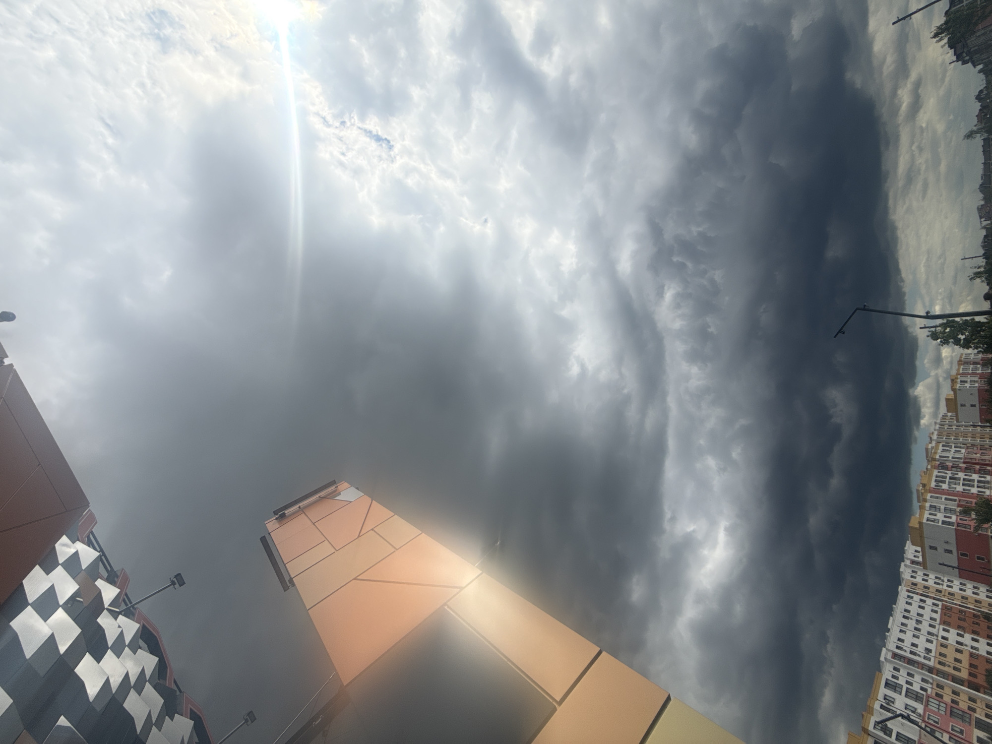 An upward-angled, close-up, full shot captures a modern, geometric building with an orange, metallic facade reflecting the bright sunlight against a dramatic sky filled with dark, heavy clouds. The building's surface is segmented into rectangular panels, with some areas illuminated by a lens flare. In the upper left, a checkered pattern of black and white squares is visible, likely part of another building or structure. Below, a panoramic view of a dense urban landscape unfolds with rows of multi-story apartment buildings painted in various colors, including white, orange, and red. The foreground is dominated by the ominous, grey clouds that suggest an impending storm. Two streetlights are visible in the lower portion of the frame, silhouetted against the cloudy sky. The scene is an urban environment, possibly a residential area, under a threatening sky.