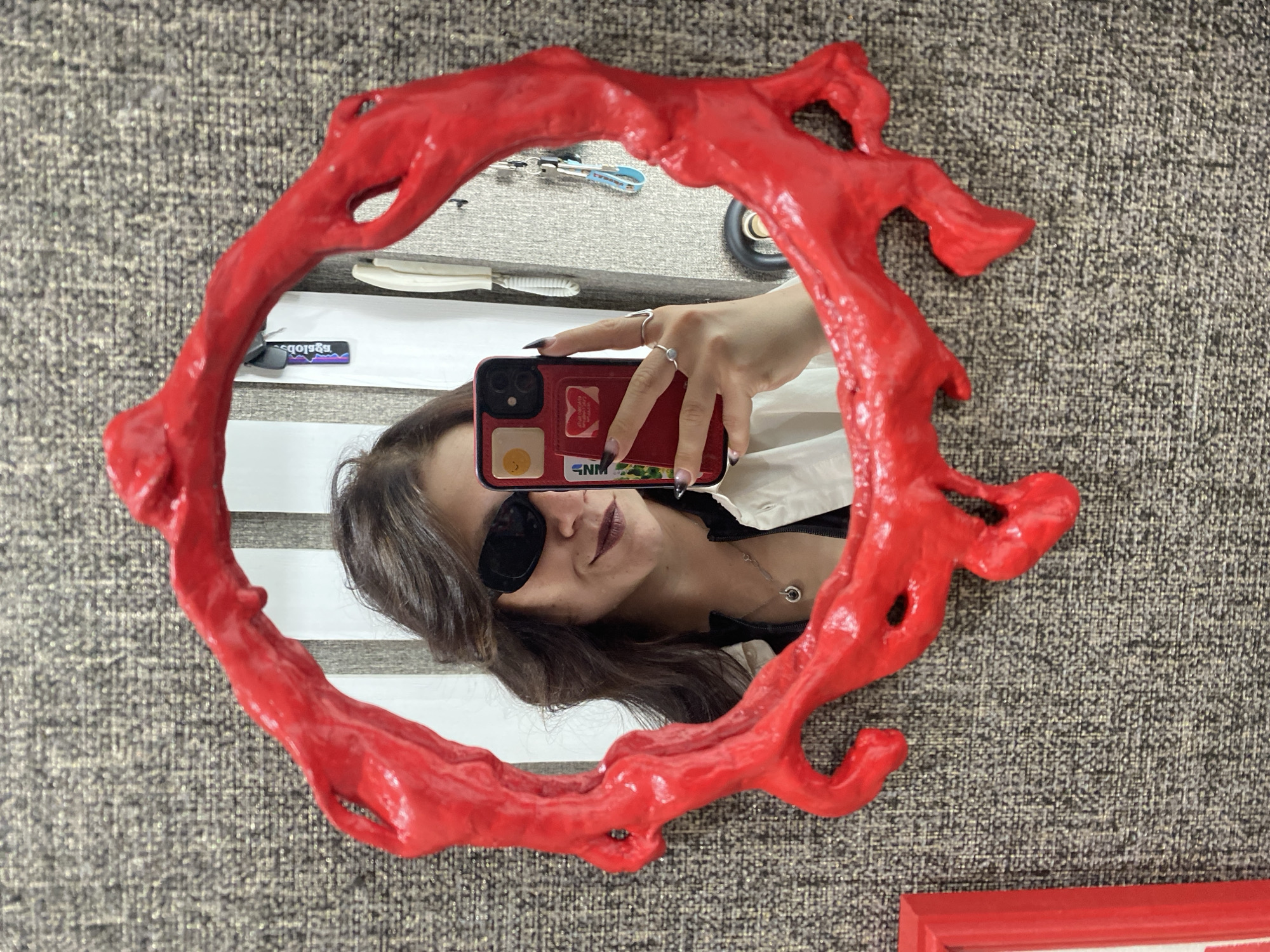 A woman takes a mirror selfie in a room. She is wearing sunglasses and a white shirt. The mirror has a red, organic-shaped frame. Behind her, a wall with a textured pattern is visible. A phone with stickers on it is held by the woman, reflecting in the mirror. The room appears to be a domestic setting. There is no identifiable information about the city of Chaikovsky, Russia in the image.