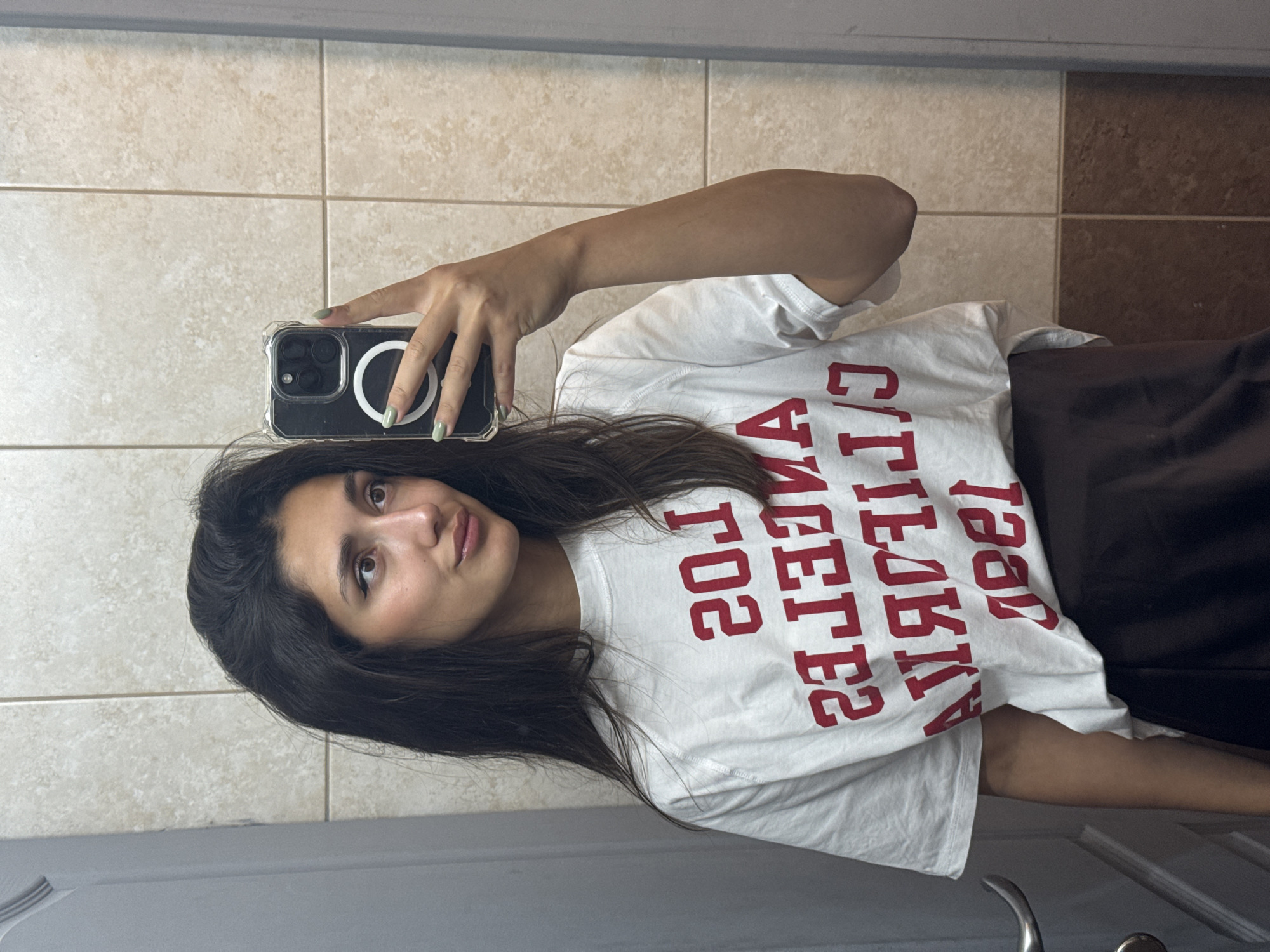 A young adult female is depicted taking a mirror selfie indoors. She has dark, long hair, brown eyes with subtle eyeliner, and light-colored lip gloss. Her right hand, with nails painted light green, holds a dark-colored smartphone encased in a clear protective cover, featuring a white circular MagSafe accessory on its rear. The phone's camera array is visible. She wears a white short-sleeved t-shirt with mirrored red text, which appears to read "LOS ANGELES CALIFORNIA". A dark brown or black skirt or lower garment is visible from her waist downwards. The background consists of light beige rectangular wall tiles with light grout in the upper section, and darker, reddish-brown rectangular wall tiles in the lower section, separated by a horizontal grout line. A silver-colored door handle is visible on the left side of the frame, and a light-colored door frame or wall section is present on the right. The lighting is even, suggesting internal illumination. This scene is reported to be located in the city of Bar, Montenegro.