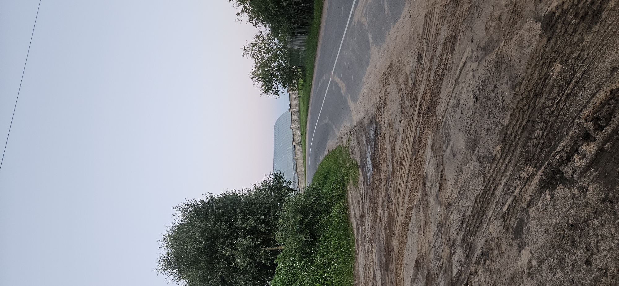 The image shows a rural road in Мазалаўскі сельскі Савет, Belarus. In the foreground, there is a dirt shoulder with tire tracks and some puddles. The road itself is asphalt with a white line marking the lane. To the left of the road, there is a dense patch of green foliage and trees. In the background, across the road, there is a long, curved structure with a translucent roof, possibly a greenhouse or agricultural building, enclosed by a stone wall and a wire fence. To the right of the road, there are more trees and a rustic wooden fence. The sky is a pale blue, suggesting either early morning or late evening. There are no people or animals visible in the image.