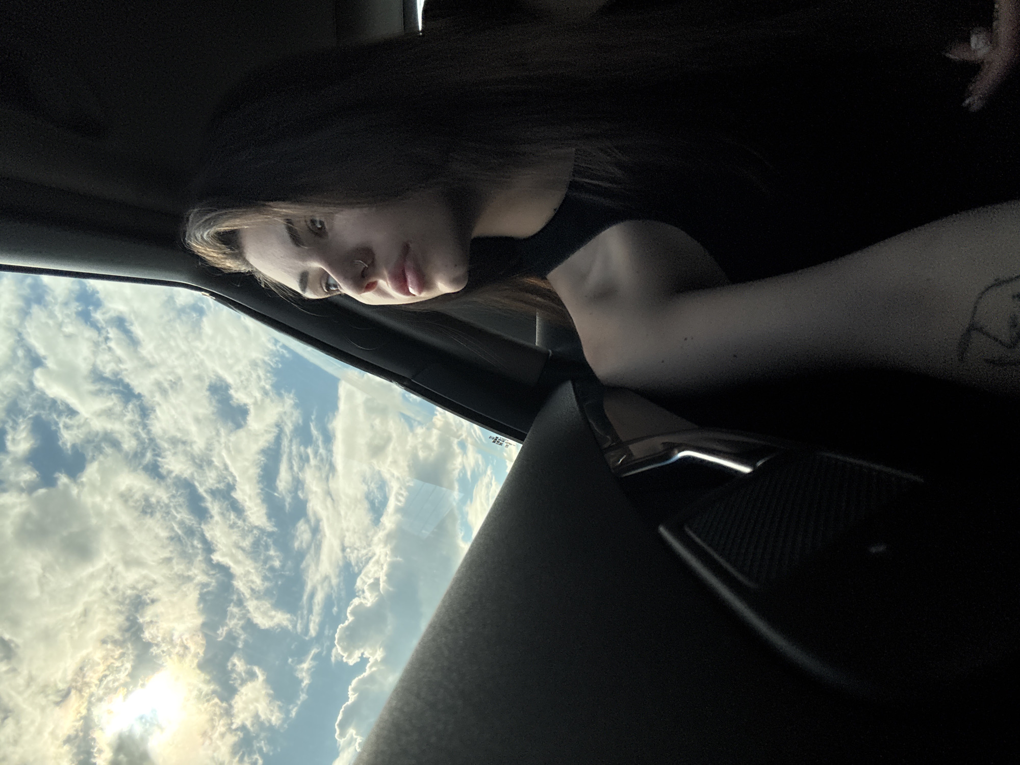 An indoor shot taken from a tilted perspective shows a young woman inside a vehicle, gazing out of a side window at a cloudy sky. The woman, with long dark hair, is positioned on the upper right side of the frame, her face turned towards the window. She wears a dark top, and a nose piercing is visible on her right nostril. Her expression appears neutral or contemplative. A tattoo is partially visible on her upper left arm.
Through the vehicle's window, which occupies the lower left portion of the image, a bright daytime sky filled with various white and grey clouds is visible. Sunlight shines brightly through a break in the clouds, creating a glare and illuminating the cloud formations with warm tones, suggesting a late afternoon or early evening setting. Parts of the vehicle's dark interior, including the headliner, window frame, and a door panel with a speaker grille and handle, frame the scene. No specific objects or activities outside the vehicle are discernible beyond the sky. No text is visible. The scene is set in or around Herakovo, Bulgaria.