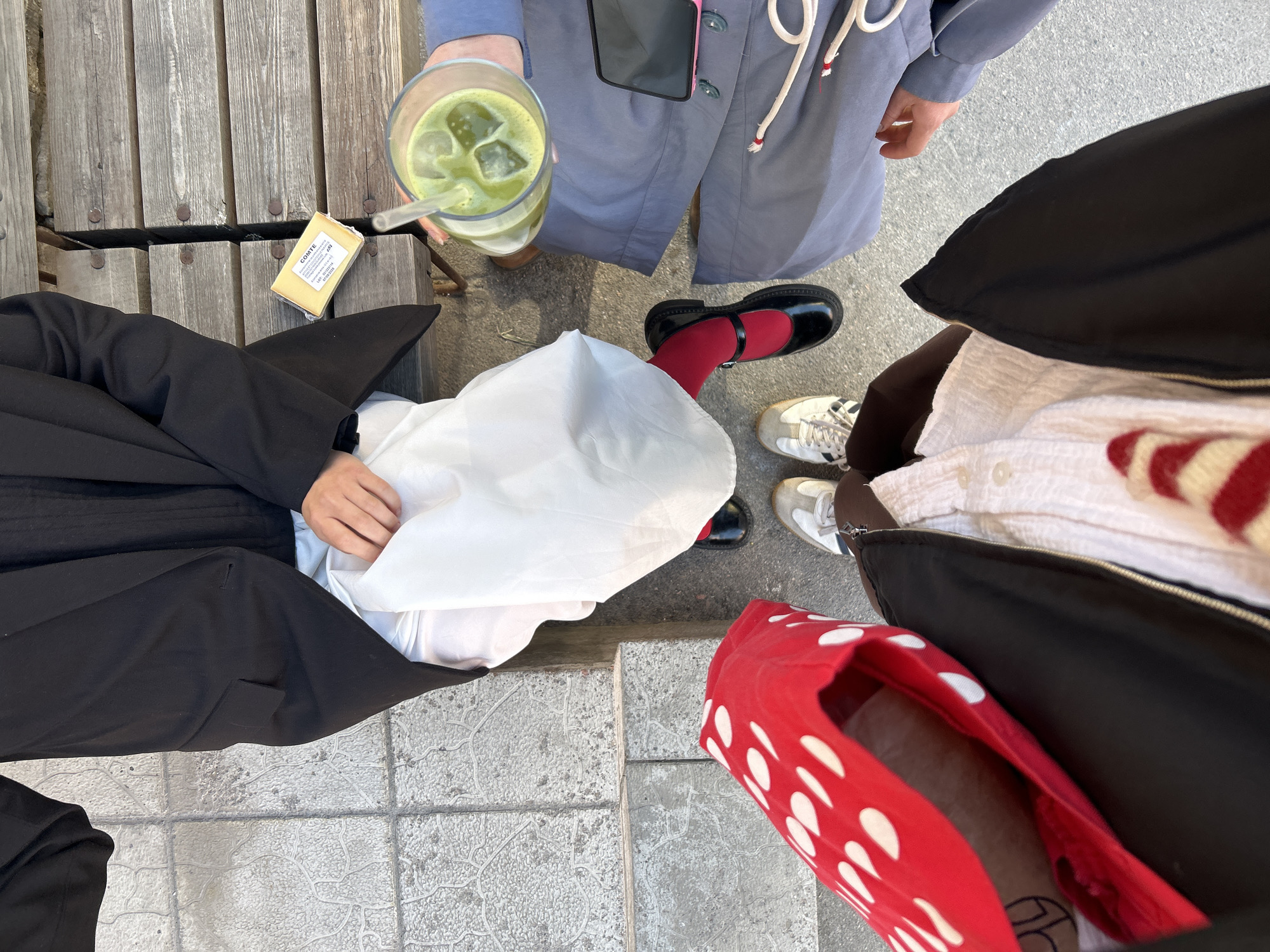 A high-angle shot shows four people standing in a public space. The person on the top left is sitting on a wooden bench and wearing a black coat over a black top. To their left on the bench is a package of Comté cheese. The person to the upper right wears a light blue coat and holds a glass of green iced drink. The person diagonally across from the green drink is wearing black outerwear and white tennis shoes. The last person on the bottom right is wearing a red and white polka dot shoe. The background consists of a grey concrete surface. The shot appears to be taken outdoors in Saint Petersburg, Russia.