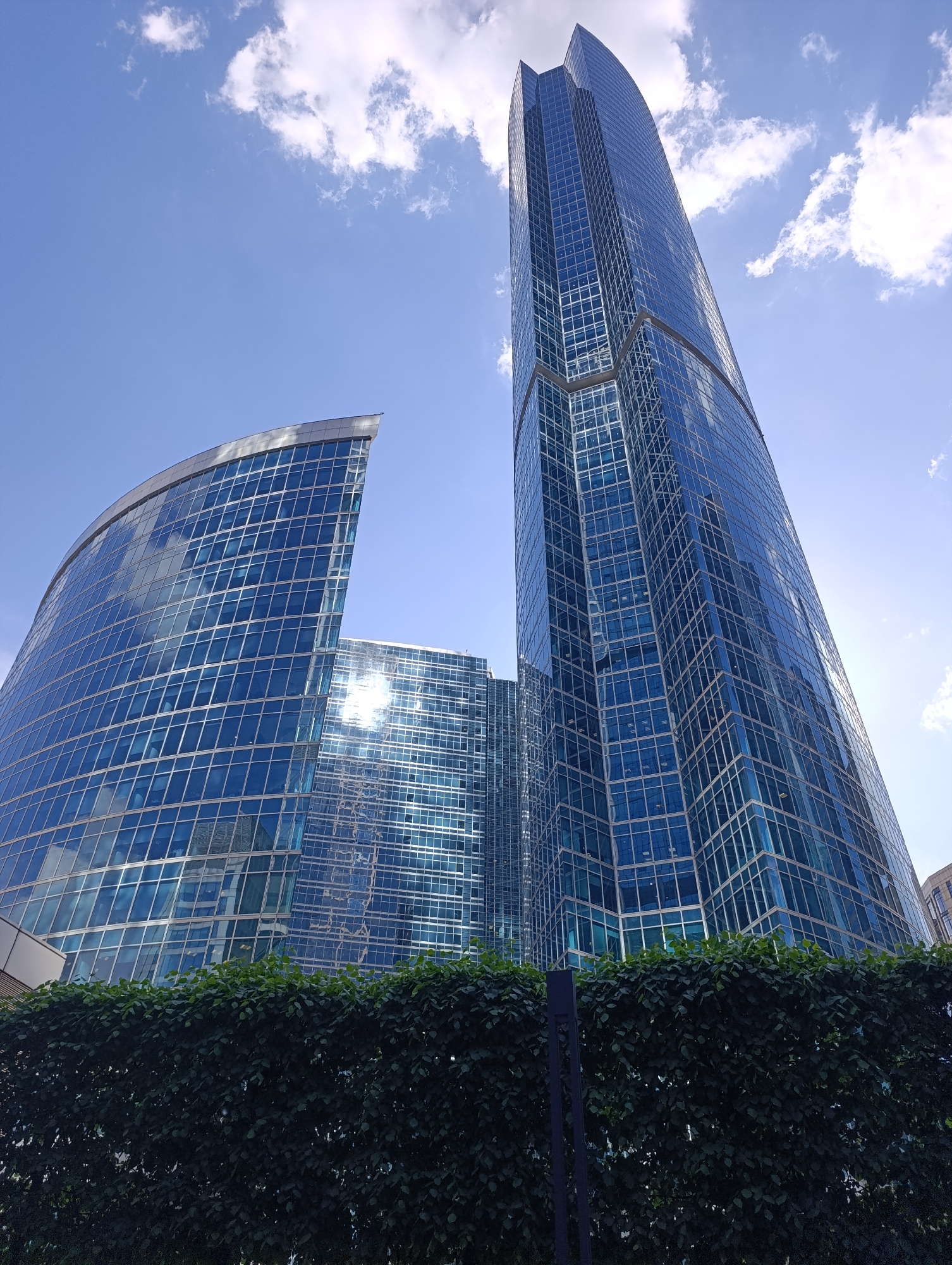 The image displays a vibrant urban scene in Moscow, Russia, centered on modern skyscrapers under a bright blue sky dotted with scattered white cumulus clouds. In the foreground, a dense, dark green hedge forms a natural barrier, extending across the bottom of the frame and partially obscuring the lower sections of the buildings. A slender, dark vertical pole is visible within the hedge on the right side.
The primary subjects are two distinct skyscrapers, both featuring expansive blue glass facades that reflect the sky. On the right, a very tall and slender building rises dramatically, its glass exterior composed of a grid of windows. Its top half tapers gently, contributing to its imposing height. To the left, a second, shorter building exhibits a striking, crescent-shaped, curved glass facade. Behind this curved structure, a third building with a similar blue glass exterior intensely reflects sunlight, creating a bright, almost star-like glare.
The scene depicts a clear and sunny day, indicated by the strong light, bright sky, and distinct reflections on the glass surfaces. The architectural style points to a contemporary business or financial district, strongly aligning with the Moscow City complex. No people or specific activities are visible, placing sole emphasis on the grandeur and design of the modern architecture. There is no discernible text within the image.