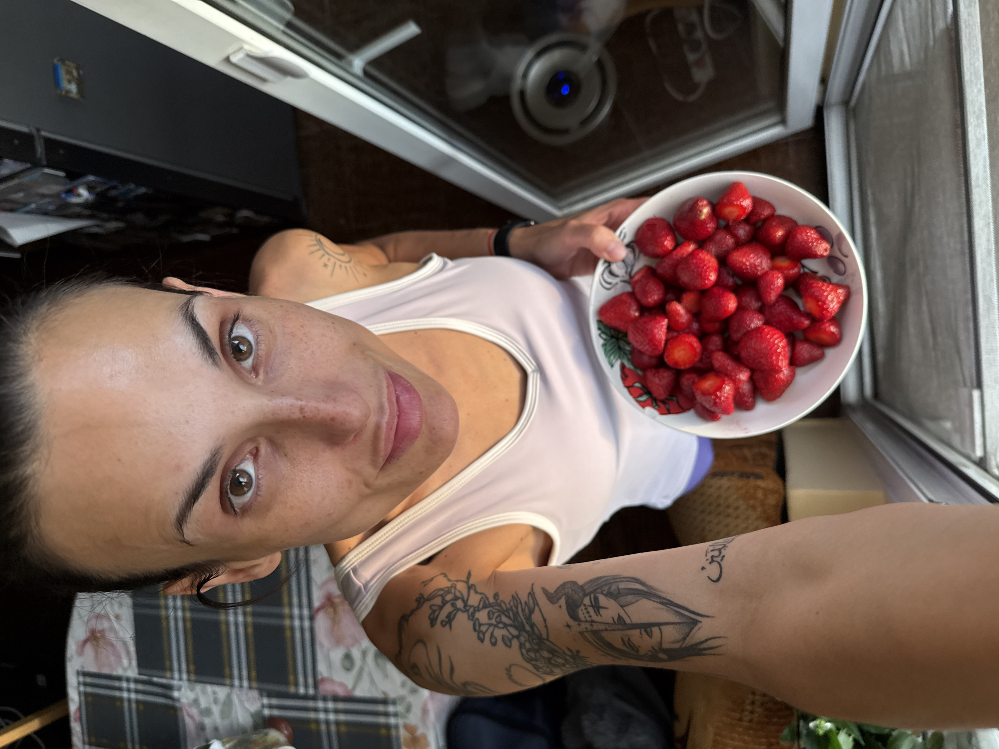 A woman is holding a bowl of strawberries. The woman is indoors, near a window. The background includes a table with a plaid pattern, and possibly some plants. The specific location in the city of Pleven, Bulgaria, is not discernible from the image.