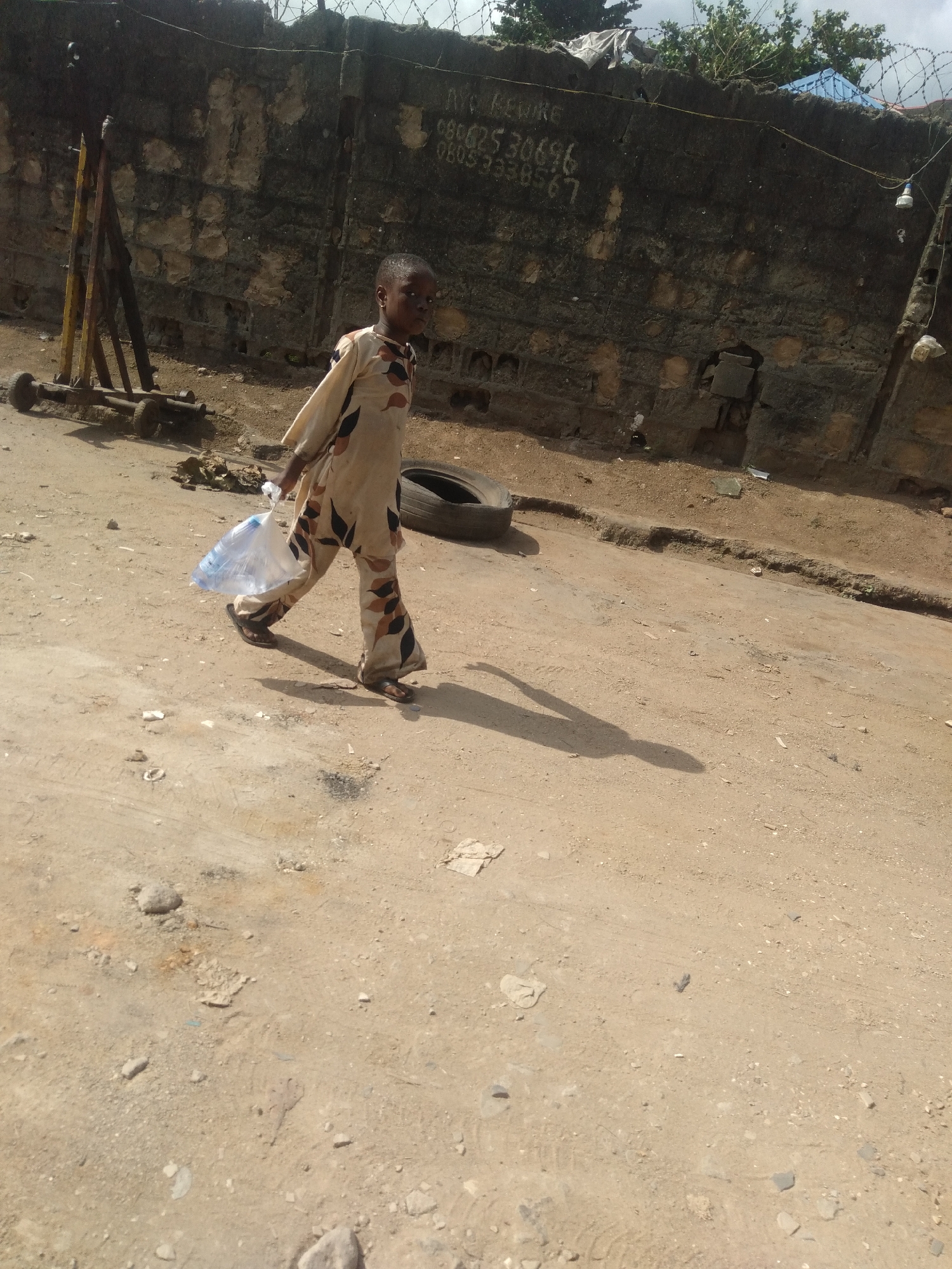 A young child is walking on a dirt road in an urban environment in the city of Unknown, Unknown. The child is wearing a patterned garment and sandals and carrying a plastic bag. Behind the child is a weathered, barbed-wire topped wall with some text written on it. There is debris and a tire to the side of the road. There is some metal scaffolding and rollers visible. The weather seems bright and sunny, casting a dark shadow of the child on the sandy soil.