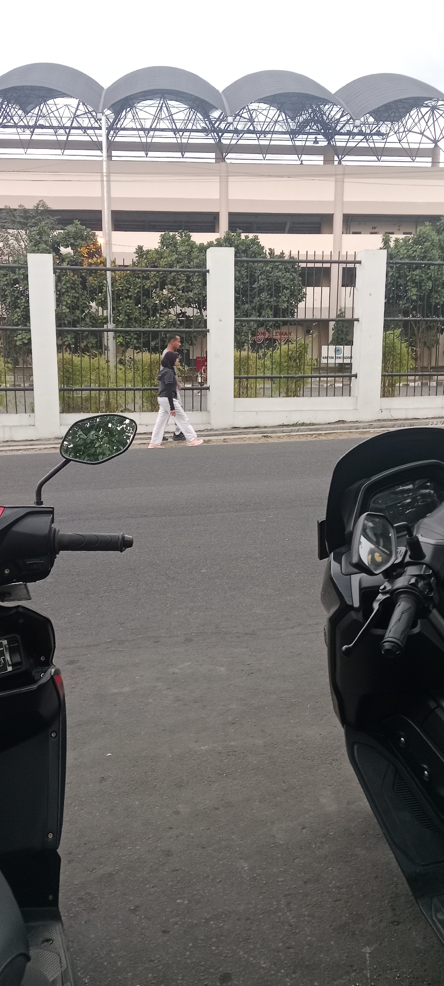 A man and a woman are walking on a sidewalk in front of a fence and a building with a large, arched roof structure in Ngemplak, Indonesia. The man is wearing a dark shirt and white pants, while the woman is wearing a dark jacket and white pants. They are walking towards the right side of the frame. In the foreground, two motorcycles are parked on the street. The motorcycle on the left is partially visible, showing its handle and part of its body. The motorcycle on the right is also partially visible, showing its handle, mirror, and a portion of its body. The street is paved, and the sidewalk is made of concrete. Behind the fence, there are trees and the building. The sky is overcast.