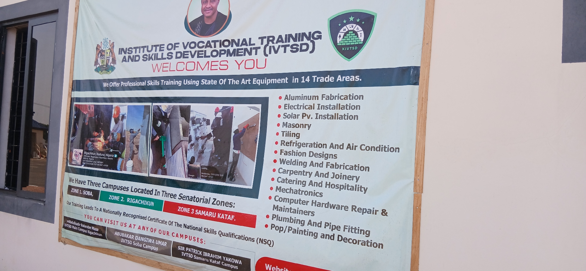 The image displays a large informational banner for the "INSTITUTE OF VOCATIONAL TRAINING AND SKILLS DEVELOPMENT (IVTSD)" affixed to the exterior wall of a building. A dark-paned window with metal bars is visible to the left of the banner.
The banner features a portrait of a man, likely a key figure, and two prominent logos: one resembling the Kaduna State coat of arms and another green, shield-like emblem with "KIVTSD" and various symbols representing different trades. The main title "INSTITUTE OF VOCATIONAL TRAINING AND SKILLS DEVELOPMENT (IVTSD)" is followed by "WELCOMES YOU".
The text advertises "Professional Skills Training Using State Of The Art Equipment in 14 Trade Areas." A comprehensive bulleted list details these areas: Aluminum Fabrication, Electrical Installation, Solar Pv. Installation, Masonry, Tiling, Refrigeration And Air Condition, Fashion Designs, Welding And Fabrication, Carpentry And Joinery, Catering And Hospitality, Mechatronics, Computer Hardware Repair & Maintainers, Plumbing And Pipe Fitting, and Pop/Painting and Decoration.
Below this, several small photographs show individuals engaged in practical training activities. One of these embedded images contains a Google Maps-like overlay displaying "Rigachikun, Kaduna, Nigeria," geographic coordinates, and a timestamp of "Wednesday, 21/01/2026 12:41 PM GMT+01:03."
The banner further indicates that IVTSD has "Three Campuses Located In Three Senatorial Zones: ZONE 1. SOBA, ZONE 2. RIGACHIKUN, ZONE 3 SAMARU KATAF." It assures that "Our Training Leads To A Nationally Recognised Certificate Of The National Skills Qualifications (NSQ)." Contact information and campus names, including "Abdulkadir Balarabe Musa IVTSD Main Campus Rigachikun," "ABUBAKAR DANGIWA UMAR IVTSD Soba Campus," and "SIR PATRICK IBRAHIM YAKOWA IVTSD Samaru Kataf Campus," are listed at the bottom, alongside a partially visible "Websit" (Website). The scene is set at an educational or training facility in Rigachikun, Nigeria.