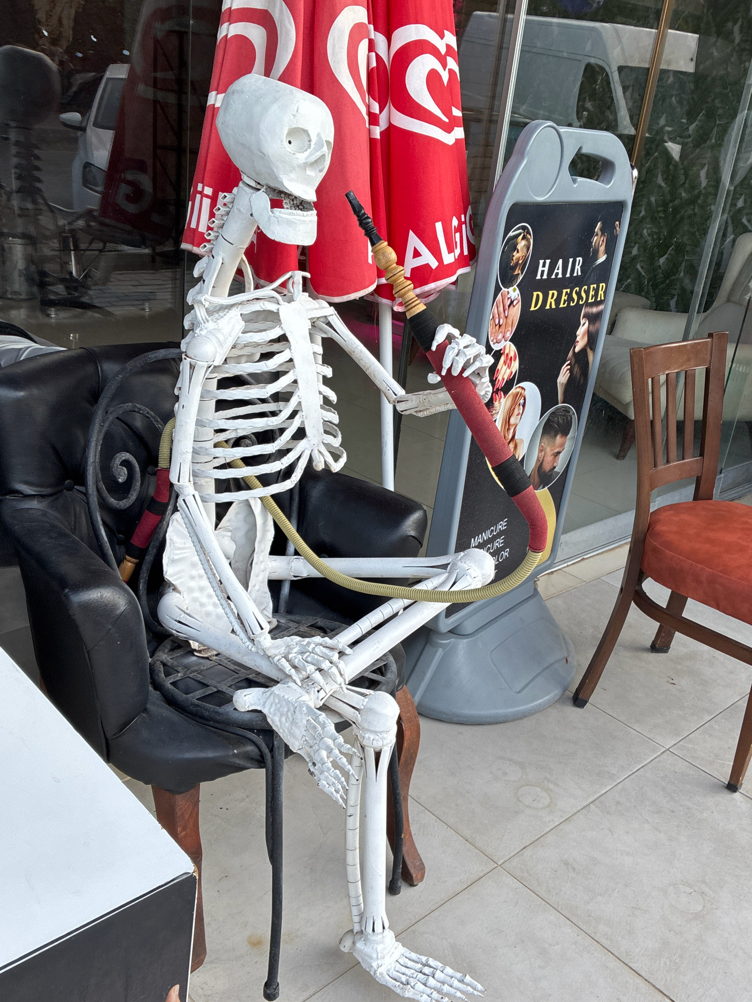 A white skeleton decoration sits in a black leather armchair outside a business in Alanya, Turkey. The skeleton is holding a hookah hose, with the mouthpiece near its mouth and the hose extending down its body. Behind the skeleton, a red umbrella with white logos is visible, as is a sign advertising "HAIR DRESSER" services, with images of hairstyles and manicures. A wooden chair with an orange seat is positioned to the right of the skeleton. The ground is tiled, and a white table is partially visible in the lower left corner of the image. The overall scene appears to be an outdoor seating area for a shop or cafe on a street in Alanya.