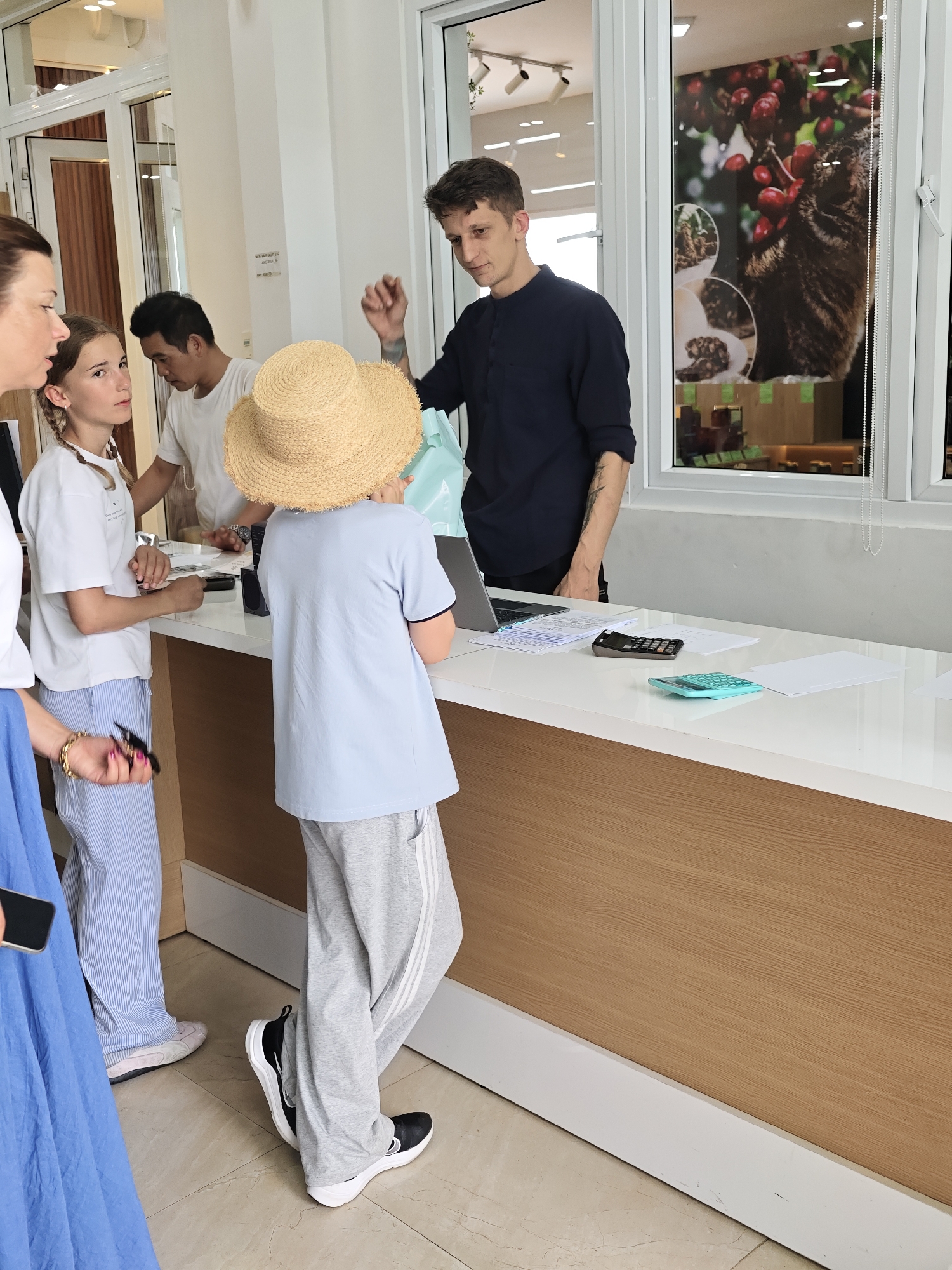 In what appears to be a cafe or shop in Lam Vien - Da Lat, Vietnam, a group of people are gathered around a counter. A man in a dark blue shirt, with tattoos visible on his arm, is standing behind the counter and gesturing with his hands. In front of him, a young person wearing a straw hat and a light blue shirt is looking towards the man. To the left of the young person, a girl with braided hair and a white t-shirt is also looking towards the counter. Next to her, a woman with brown hair, wearing a blue dress, is partially visible. Another man in a white t-shirt is standing behind the counter, between the man in the blue shirt and the girl. On the counter, there are papers, a laptop, and two calculators, one black and one teal. The environment seems to be indoors, with a window in the background displaying a poster with a cat and coffee beans.
