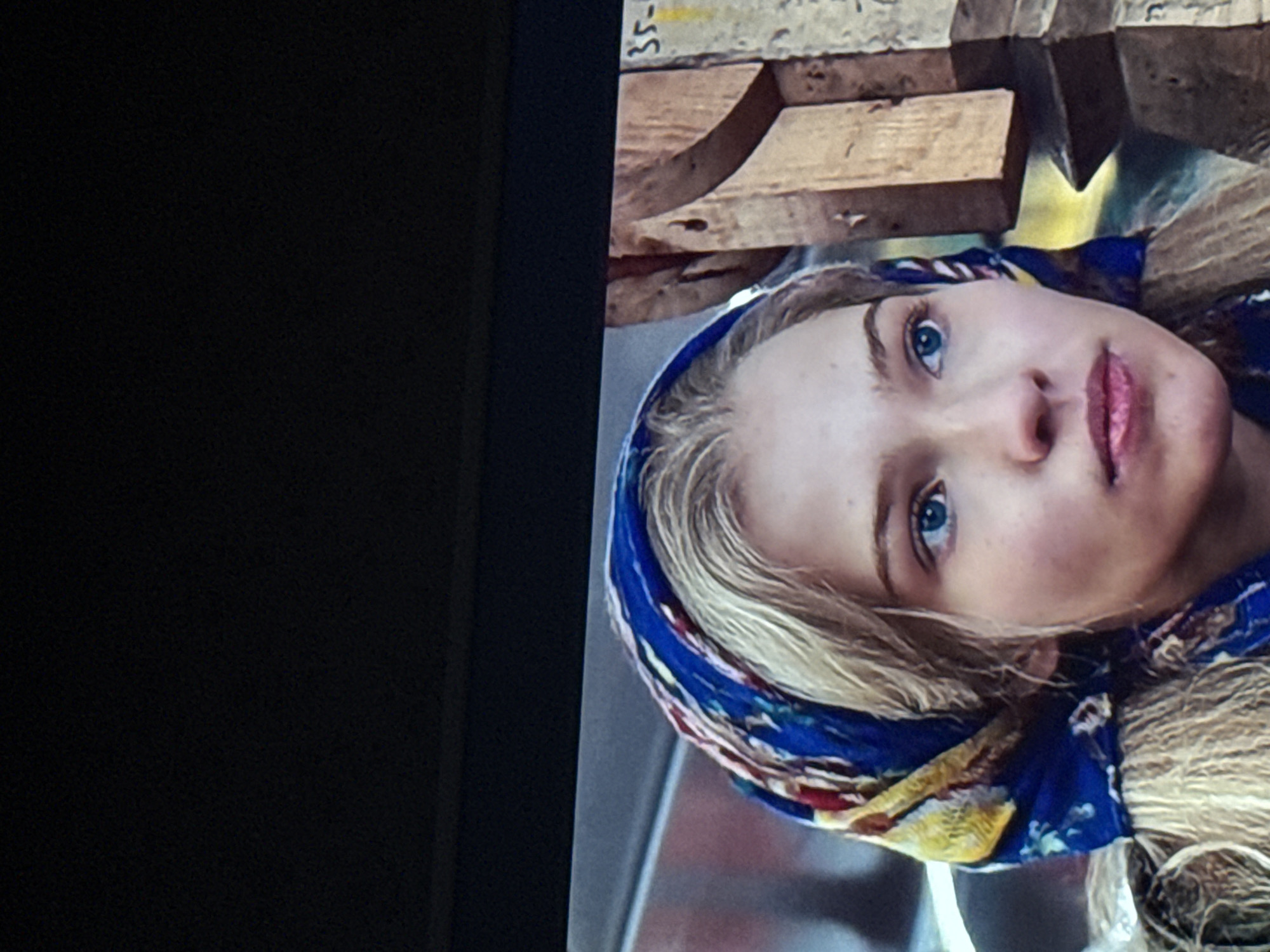 A close-up shot of a young woman with blonde hair and blue eyes wearing a blue floral headscarf. She is looking upwards. Above her are stacked wooden planks with the number "35-" visible on the top plank. The location, Mytishchi, Russia, is not explicitly visible within the image.