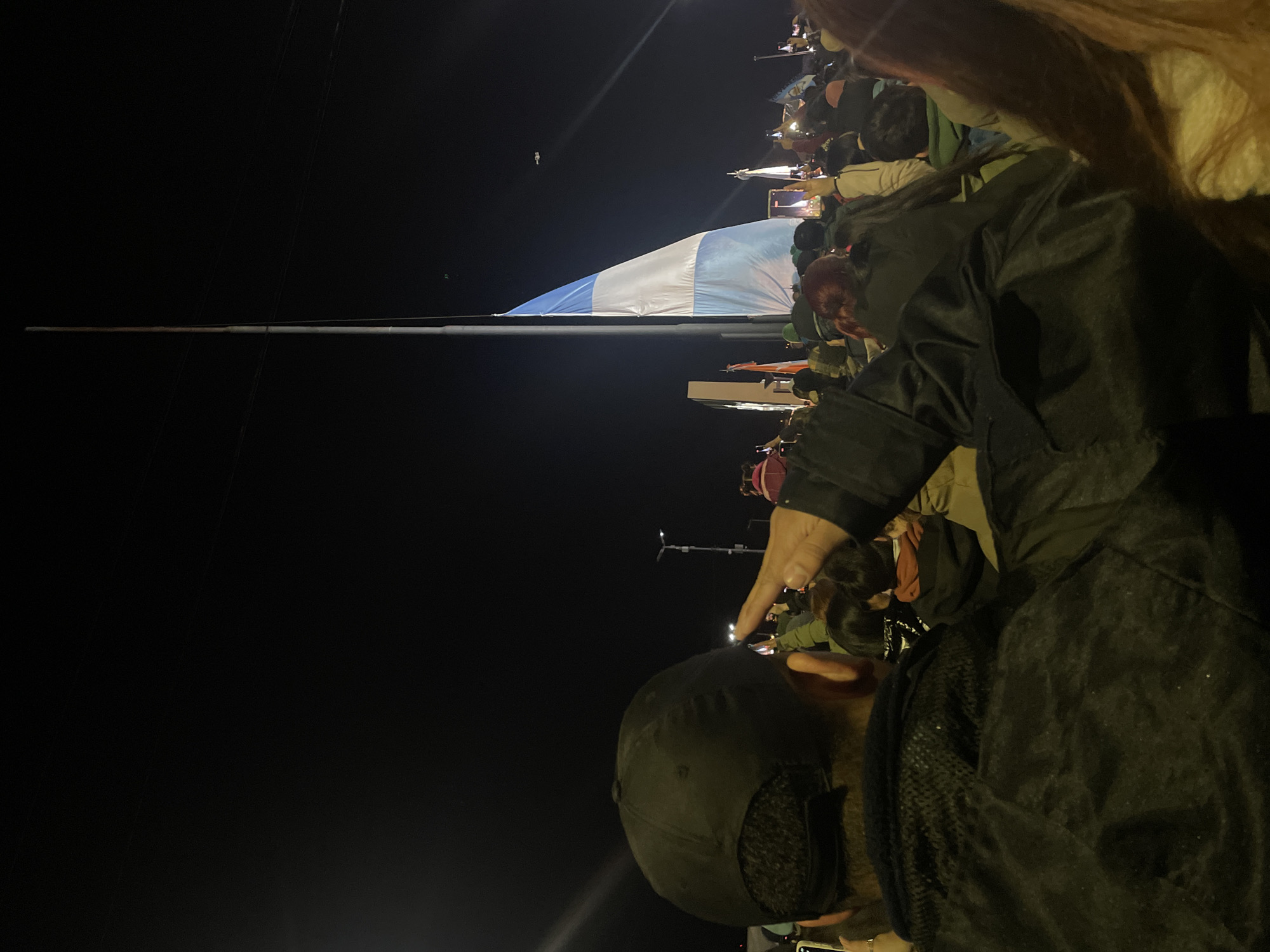 The image shows a crowd of people at night, some holding phones up to record. There is a large blue and white flag stretched horizontally above the crowd. The background is dark, with visible wires and a few lit objects. The location is likely in Ushuaia, Argentina, due to the flag's colors resembling the Argentinian flag. A person in the foreground has a hand raised, possibly pointing towards the flag.