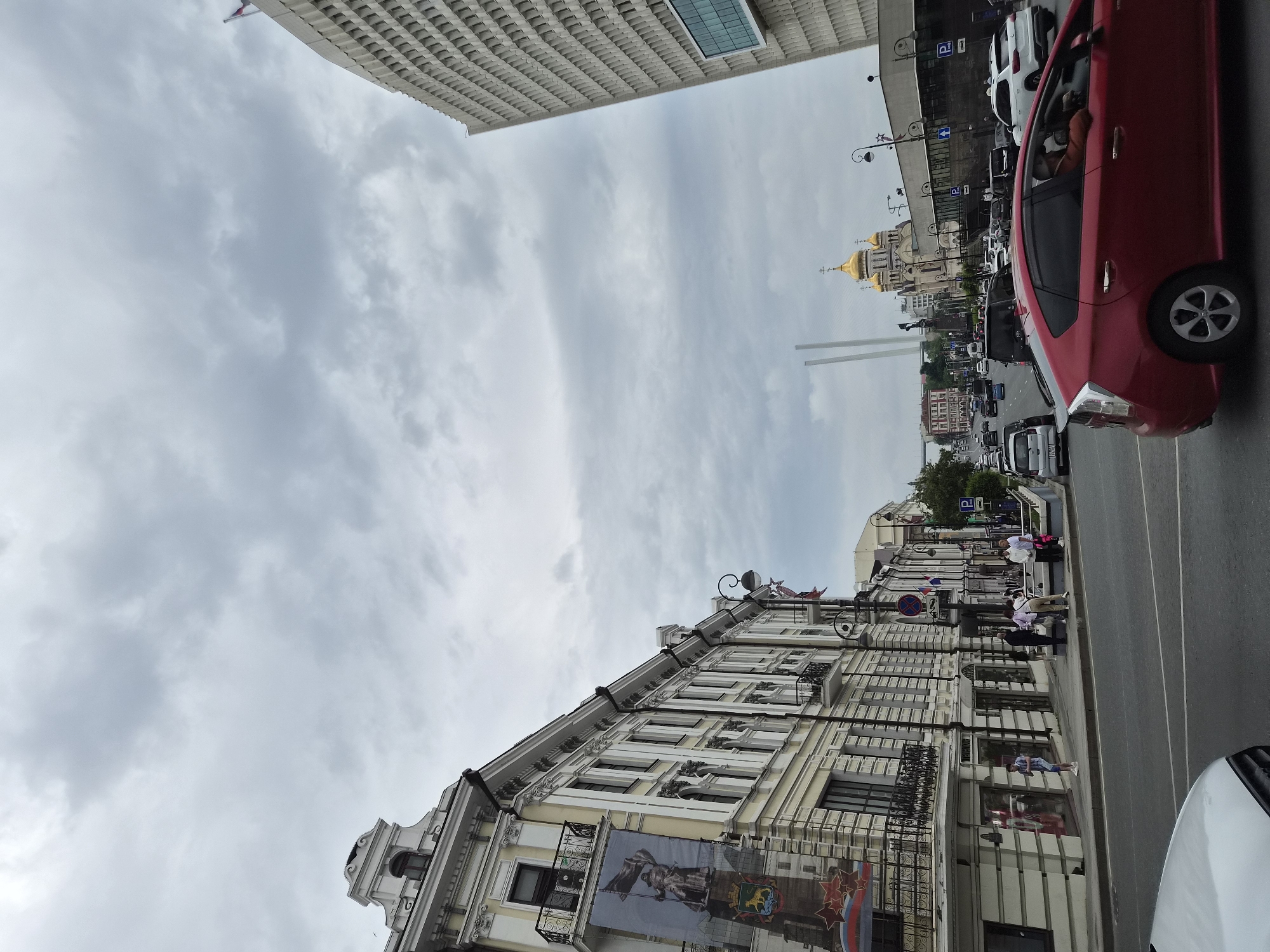 The image shows a street scene in Vladivostok, Russia, on an overcast day. In the foreground, a red car is driving on the right side of the road. To the left, a white car is parked. The street is lined with buildings. On the left side of the street, a multi-story building with ornate architecture and large banners is visible. Further down the street, there are more buildings, including a large, modern building with a grid-like facade on the right side of the image. In the distance, a golden-domed church and a bridge with tall pylons can be seen. Several people are walking on the sidewalk and some cars are driving on the road. The sky is filled with gray clouds.