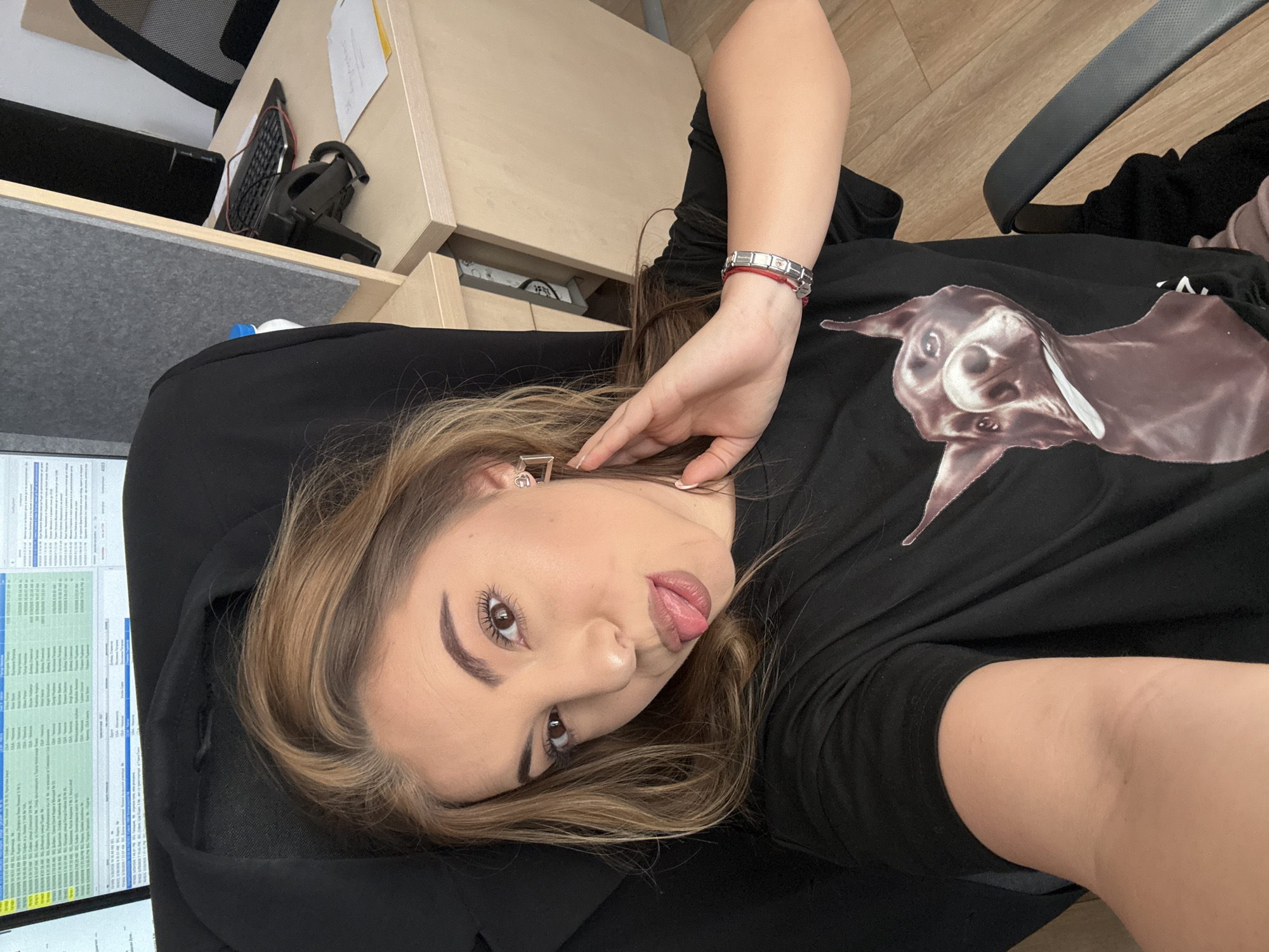 A woman is lying on a black office chair, wearing a black T-shirt with a dog's head print on it. She has brown and blonde hair and is sticking out her tongue. The woman is wearing an earring and a bracelet. Behind her is a wooden desk and computer equipment. In the background is a computer monitor, a keyboard, and an office phone. The photo seems to be taken in an office environment, and while the city of Sofia, Bulgaria is not directly identifiable from the image, it is provided as additional information in the instructions.