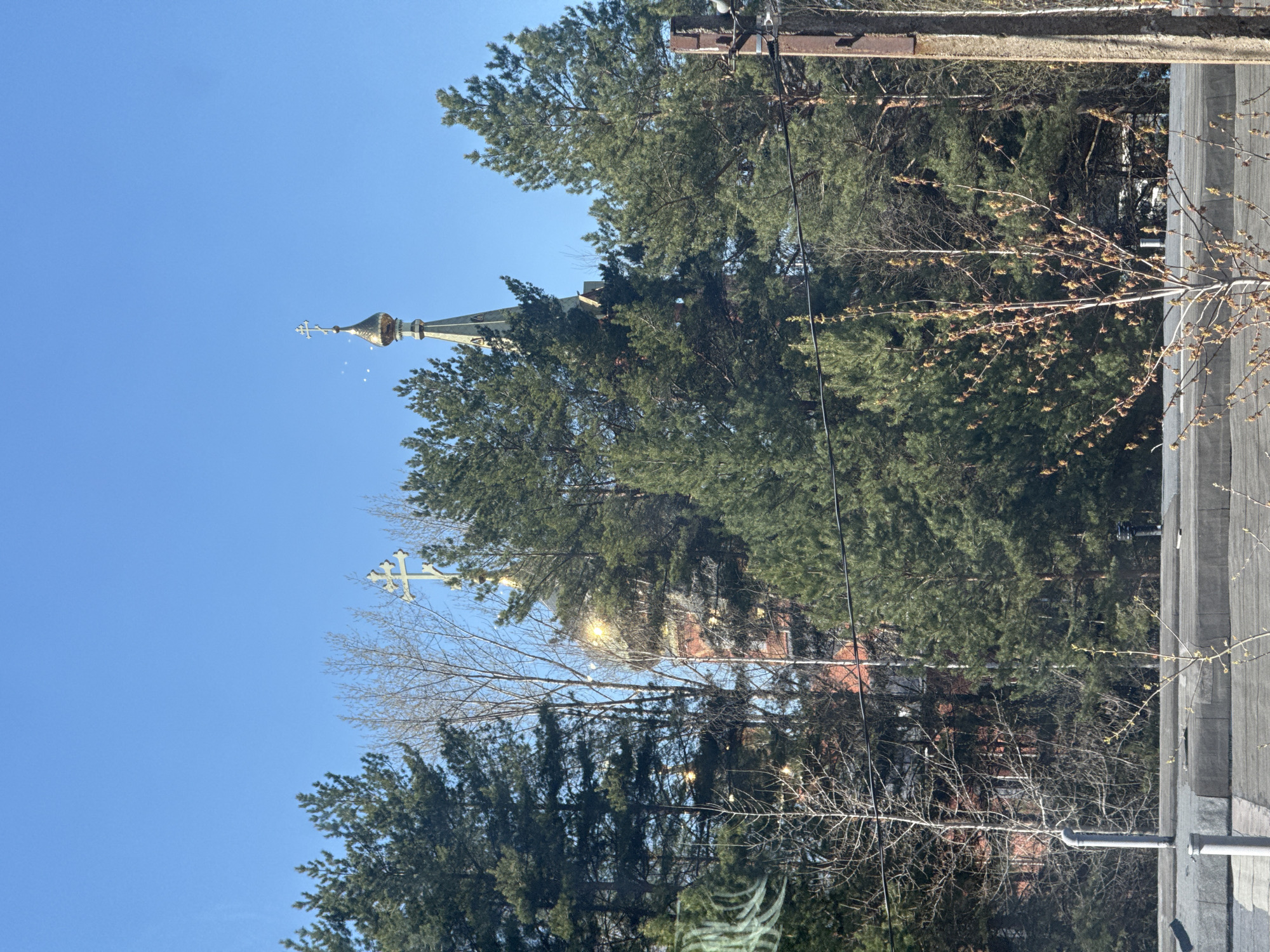 The image shows a view of a church steeple and domes emerging from behind dense evergreen trees. The sky is a clear, bright blue. A white cross stands atop one of the domes. Power lines run through the scene. The photo appears to be taken from inside a building or vehicle, with a window frame visible in the lower right corner. The location is Unknown, Unknown.