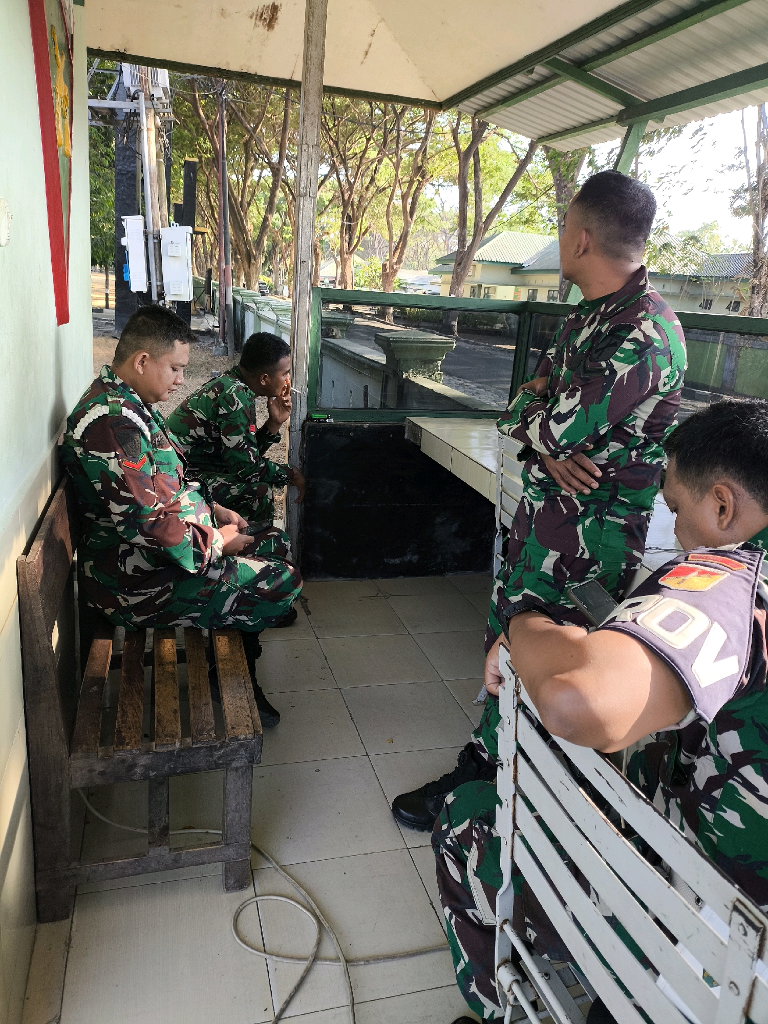 Four men in camouflage military uniforms are standing and sitting under a covered outdoor area. The men are of Asian descent and have short hair. Two men are sitting on a wooden bench to the left, one man is standing with his arms crossed, and another man is partially visible in the foreground on the right. The man second from the left is smoking a cigarette. The environment appears to be an outdoor area with a tiled floor, a wooden bench, and a white metal bench. In the background, there are trees, a building with a green roof, and what appears to be a guard post or checkpoint. The lighting suggests it is daytime, possibly late afternoon due to the shadows.