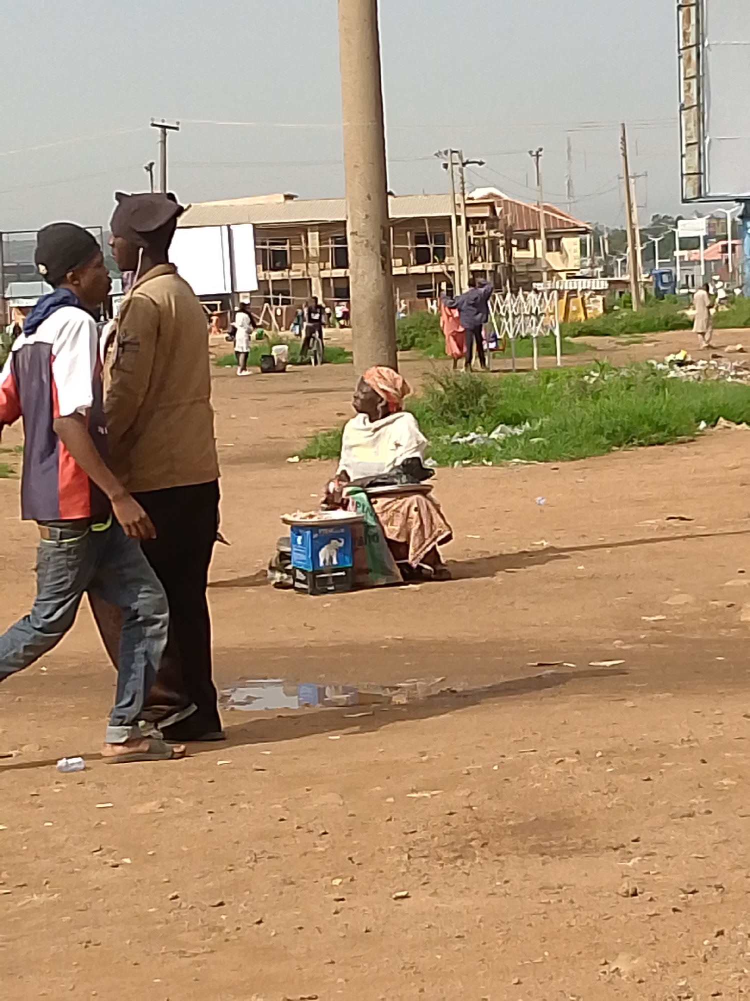 The image depicts a street scene in Bukuru, Nigeria, on a bright, possibly overcast, day. In the foreground, two young men are walking, one in a red, white, and blue jacket and jeans, the other in a tan jacket and black pants. To their right, an older woman is seated on the ground behind a small blue cart, which displays some items for sale. She is wearing a light-colored headscarf and a patterned skirt. Behind them, a dirt road stretches into the distance, lined with buildings under construction or in various states of repair. A prominent utility pole stands near the center of the frame. Other individuals are visible in the background, including some standing near a stall and a person on a bicycle. The environment appears somewhat dusty, with patches of green grass and scattered debris along the roadside. The overall impression is of a bustling urban setting with ongoing commercial activity.
