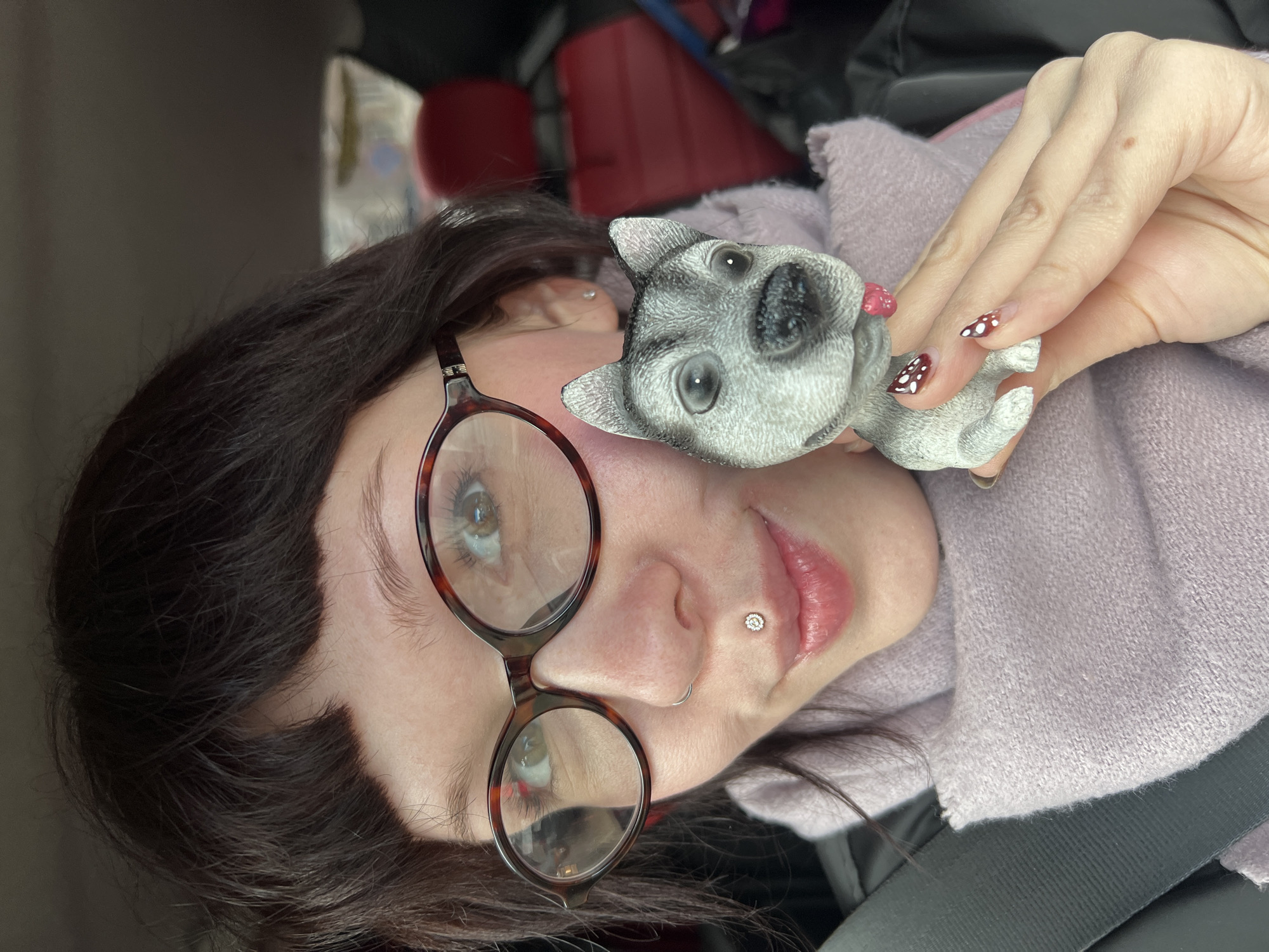 A fair-skinned woman with dark hair and round glasses is holding a small dog figurine. She has a piercing on her nose and a piercing in her ear. Her fingernails are painted with a red and white polka dot pattern. She is wearing a light purple sweater and a seatbelt is visible across her body. The background is out of focus but appears to be the inside of a car. The location, Chaikovsky, Russia, is not discernible from the image.