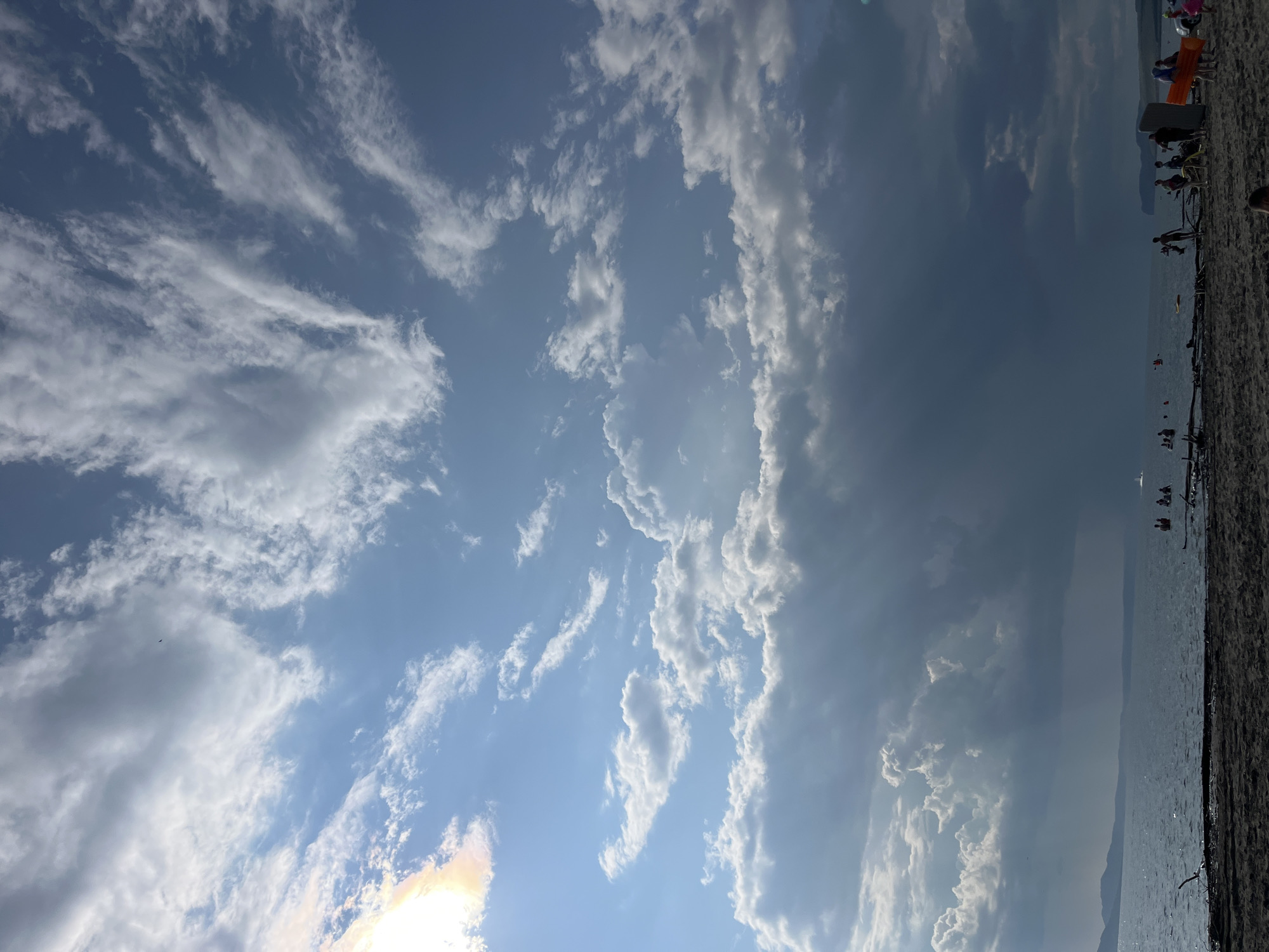 The image shows a beach scene with a body of water, likely a lake or river, in the foreground and a cloudy sky overhead. The sky is a mix of blue and dramatic dark clouds, with some shafts of sunlight breaking through. There are a few people visible on the beach and in the water, some appearing to be swimming or wading. The beach itself is composed of dark sand and some driftwood. The overall impression is of a somewhat overcast day at a recreational area. The location is not explicitly identifiable as Krasnoturansk, Russia based on visual cues alone.