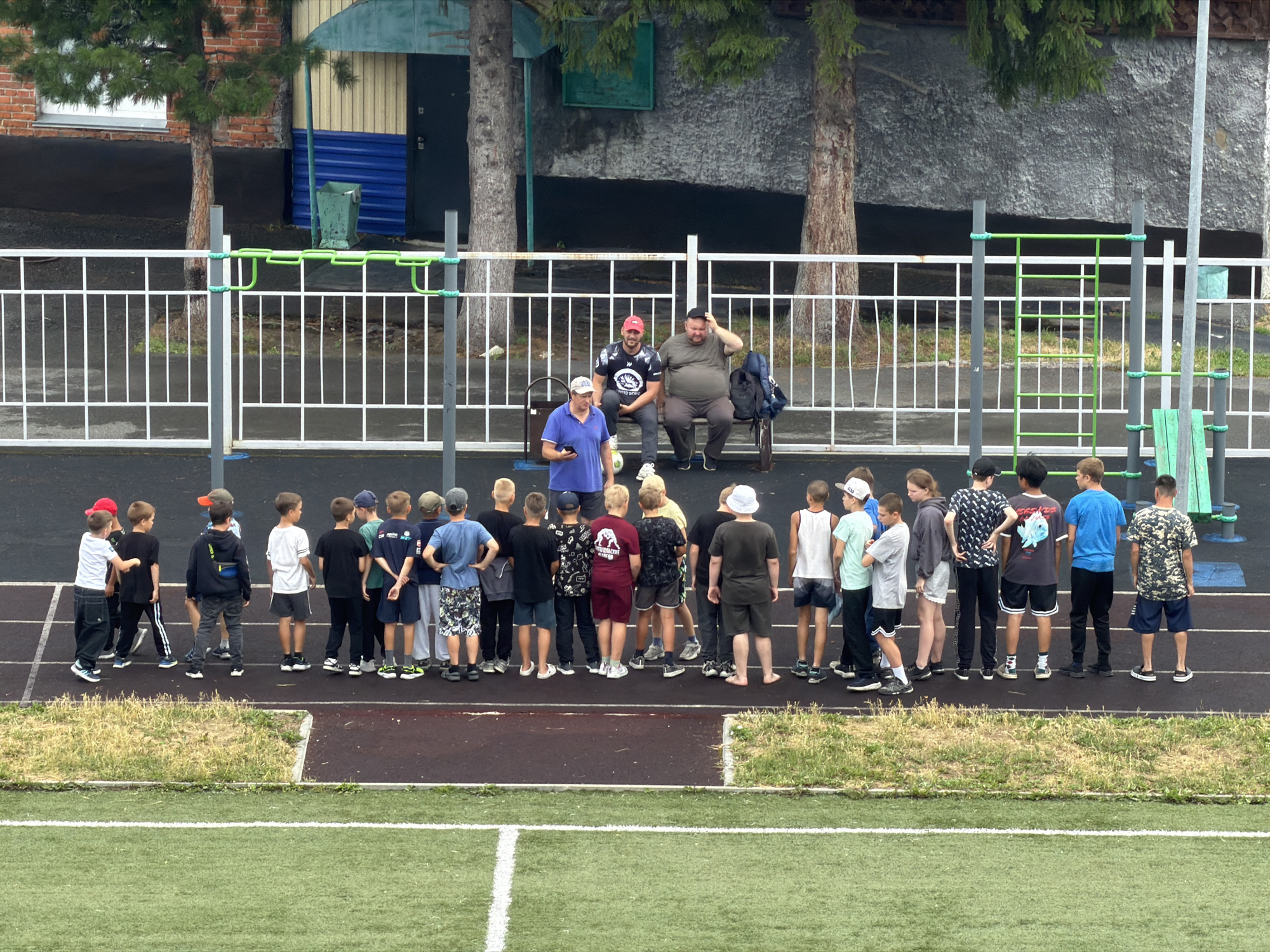 A group of children, possibly a sports team, stands on a running track facing away from the camera. They are gathered behind a white fence with green accents. Two adults, one in a red cap and a black t-shirt, the other in a gray t-shirt and dark pants, are seated on a bench to the right of the fence. A third adult in a blue t-shirt and light pants stands to the left of the fence, facing the children. The background features a building with a brick and corrugated metal exterior, along with trees. A green trash bin is visible near the building. The scene appears to be an outdoor sports facility. The image is likely taken in Tashtagol, Russia, given the context.