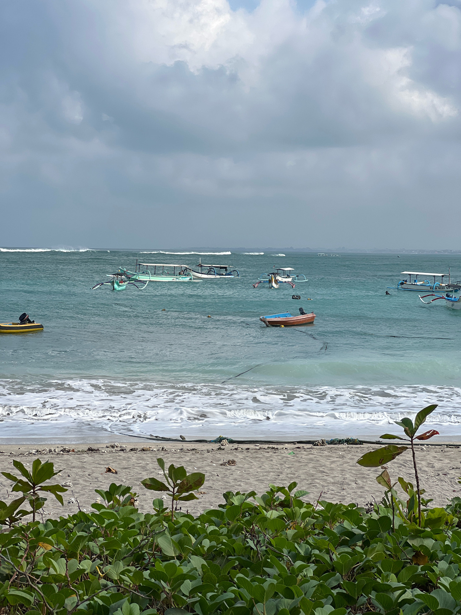The image captures a serene view of the ocean in Kuta, Indonesia. The scene is dominated by the turquoise waters, with several Balinese jukung boats scattered across the surface. The boats, characterized by their outriggers, are anchored or gently drifting in the calm sea. Some boats have canopies, suggesting they are used for fishing or transporting tourists. Waves are breaking on the shore, creating a foamy white line where the water meets the sand. The beach itself is sandy and appears somewhat weathered, with some debris and coral fragments visible. In the foreground, lush green vegetation with broad leaves grows along the edge of the beach, framing the view of the ocean. The sky is overcast with a mix of grey and white clouds, indicating a possibly cloudy or approaching stormy day. The overall impression is one of tranquility and natural beauty, characteristic of a tropical coastline.