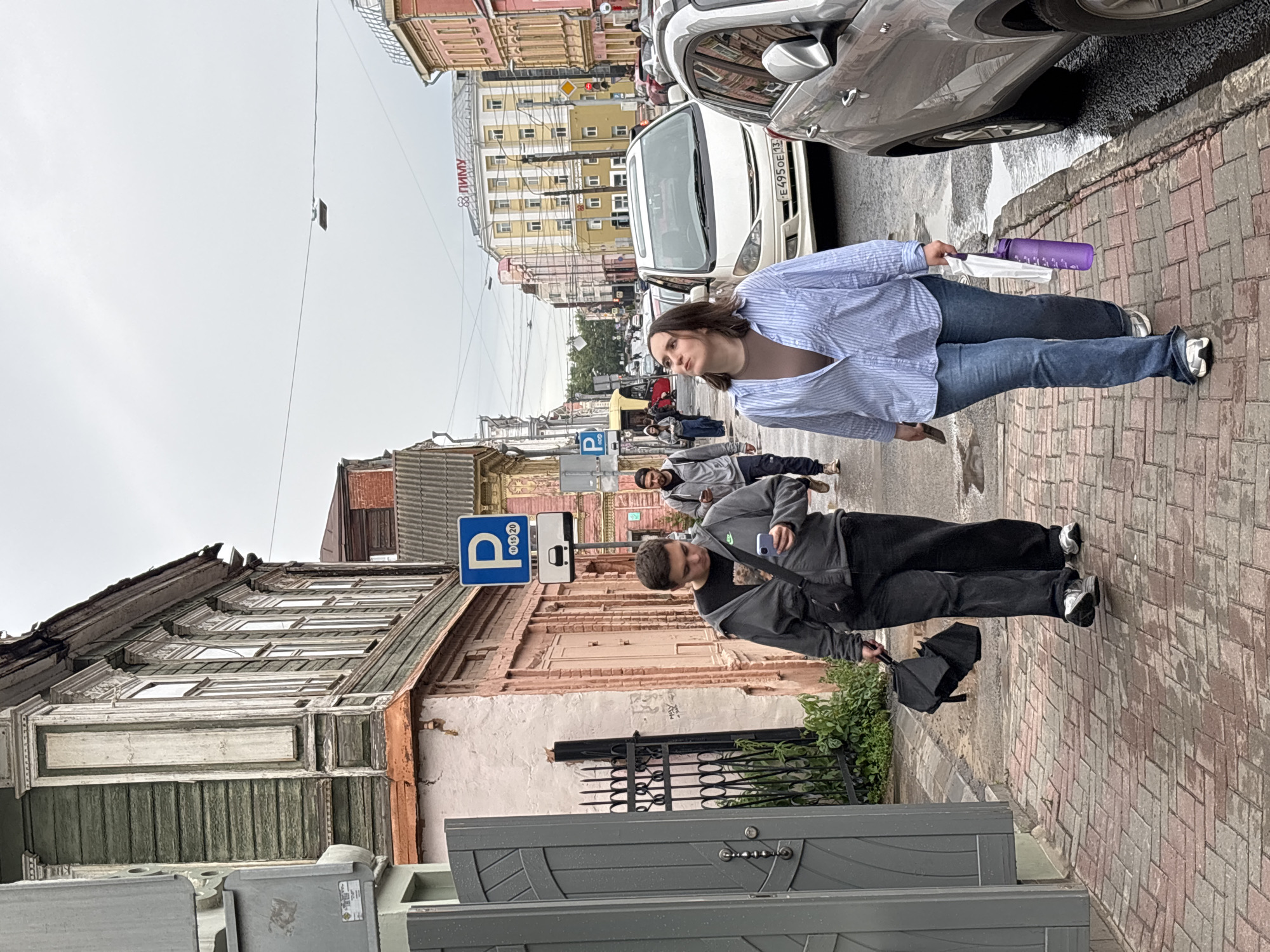 In Nizhny Novgorod, Russia, on a cloudy day, two individuals walk down a pedestrian sidewalk alongside a street with parked cars and buildings. The sidewalk is paved with red brick.
The person in the foreground on the left, a male, walks facing forward, looking down at his phone. He wears a grey hooded jacket over a dark t-shirt, dark pants, and grey sneakers. He carries a black crossbody bag and a folded black umbrella.
The person in the foreground on the right, a female, walks facing forward and slightly to the left, looking upwards. She wears a light blue striped button-up shirt, a brown undershirt, blue jeans, and white sneakers. She carries a white folded item and a purple water bottle in her right hand.
Behind them, another male walks in the same direction, wearing a grey hooded jacket and dark pants. Other pedestrians are visible further down the street, as are cars, including a white minivan and a silver SUV. The buildings lining the street vary in style and color, with some featuring ornate architectural details. A blue parking sign with white lettering is visible on the left side of the street. The street appears wet, suggesting recent rain.