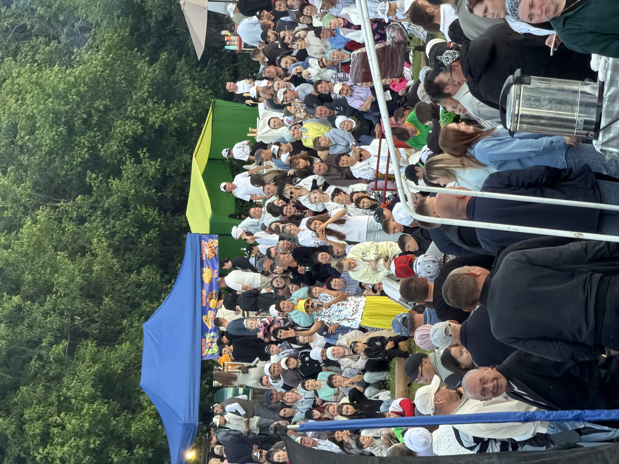 The image depicts a large outdoor gathering or festival taking place in an unknown location. A significant crowd of diverse individuals, ranging from children to elderly adults, is assembled on a grassy or dirt embankment, many seated and facing a central area. The backdrop consists of dense, green deciduous trees.
At the center of the scene, slightly elevated, a performer or speaker is visible. This individual wears a reflective silver sequined top, a bright yellow long skirt, and a yellow brimmed hat, with a dark mustache, and is holding a microphone while addressing the audience. Many audience members are observing the performer, with some smiling, and at least one person in the mid-ground capturing a photo with a mobile phone.
In the upper left quadrant, a large blue canopy tent is set up, featuring banners beneath its front edge that display images of food items such as burgers, fries, and other dishes, along with some text, likely menu descriptions. To the right of the blue tent, two lime-green canopy tents are present, with people gathered around them, suggesting food or drink service. An off-white or light brown patio umbrella is visible on the far right.
In the lower right foreground, a metal beverage urn or dispenser sits on a table covered by a white tablecloth, with several individuals standing nearby. The crowd in the foreground is primarily standing, with some individuals looking towards the camera while others face away, indicating a dynamic gathering. Metal railings are discernible in the lower foreground, possibly part of a barrier or seating structure. The overall lighting suggests daytime, possibly late afternoon, with diffused light illuminating the scene.